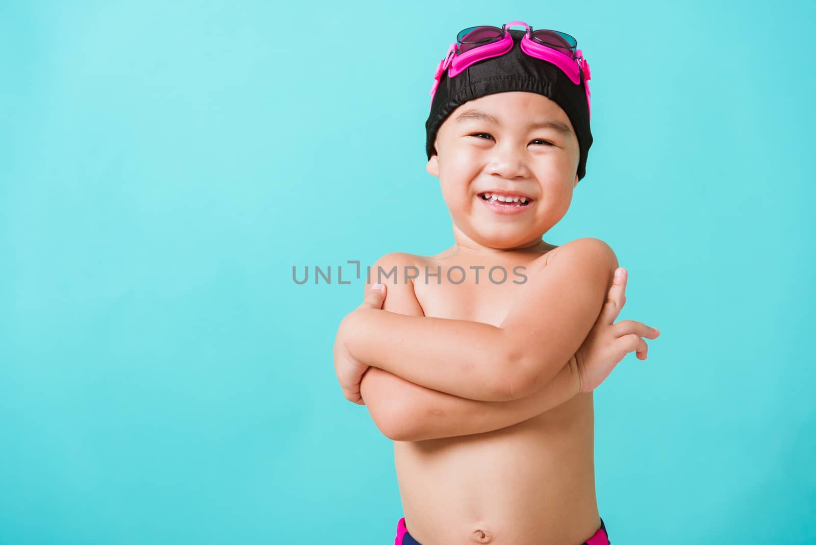 Summer vacation concept, Closeup portrait Asian happy cute little child boy wear goggles and swimsuit, Kid having fun with in summer vacation crossed arm, studio shot isolated blue background