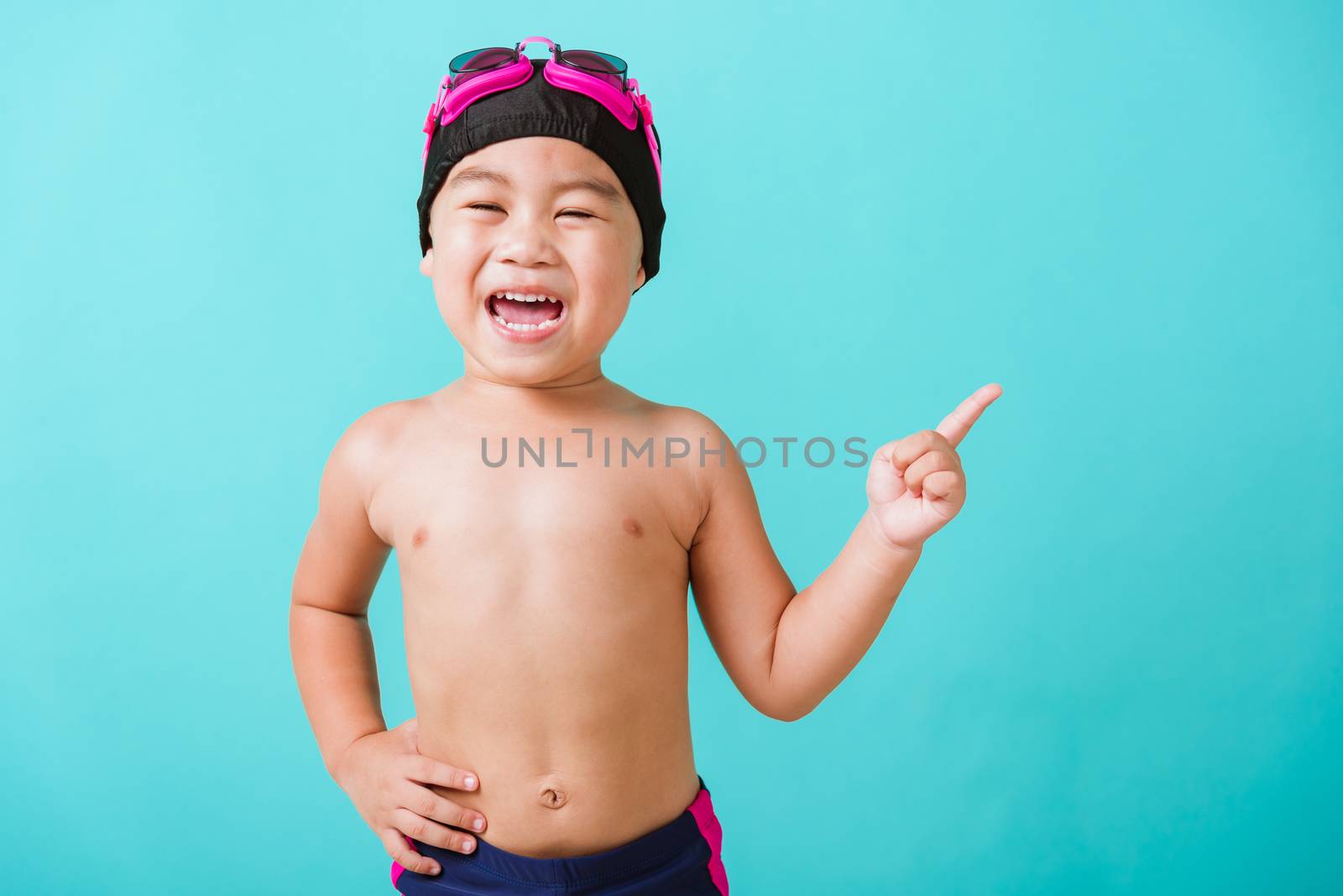 Summer vacation concept, Closeup portrait Asian happy cute little child boy wear goggles and swimsuit, Kid having fun with in summer vacation point to side away, studio shot isolated blue background