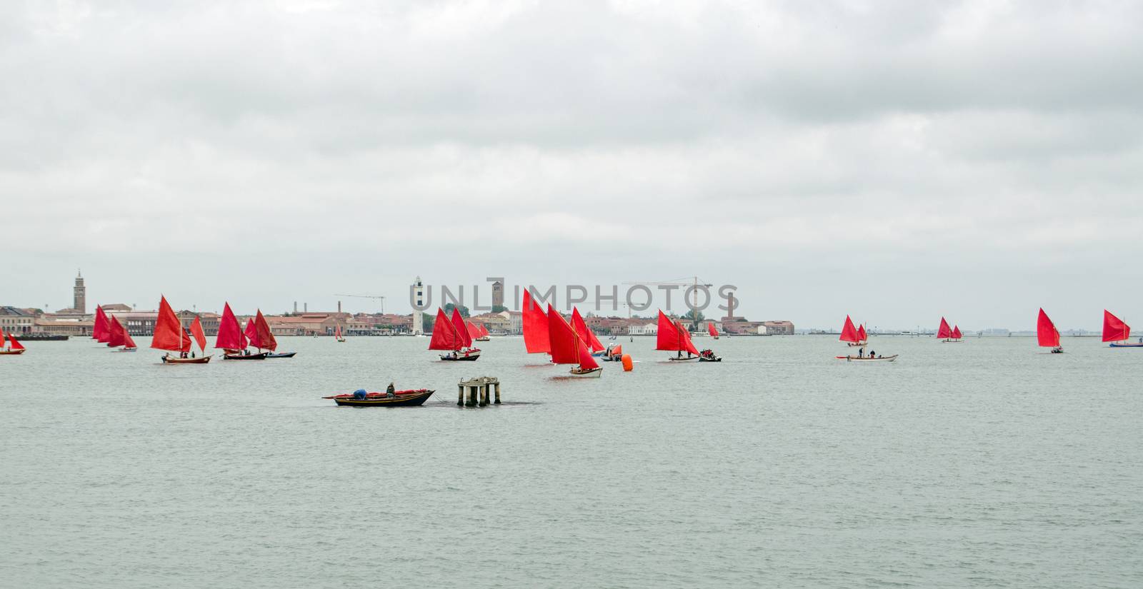 A regatta of Venetian sailing boats, all sporting red painted sails, on the lagoon near Murano.  Part of the Red Regatta event organised by artist Melissa McGill on an overcast afternoon in May, Venice.
