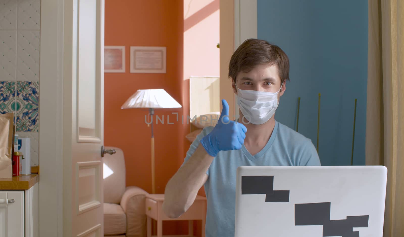 Young man showing Thumb up. He is working from home on computer in mask and blue rubber gloves. Quarantine, coronavirus epidemic.