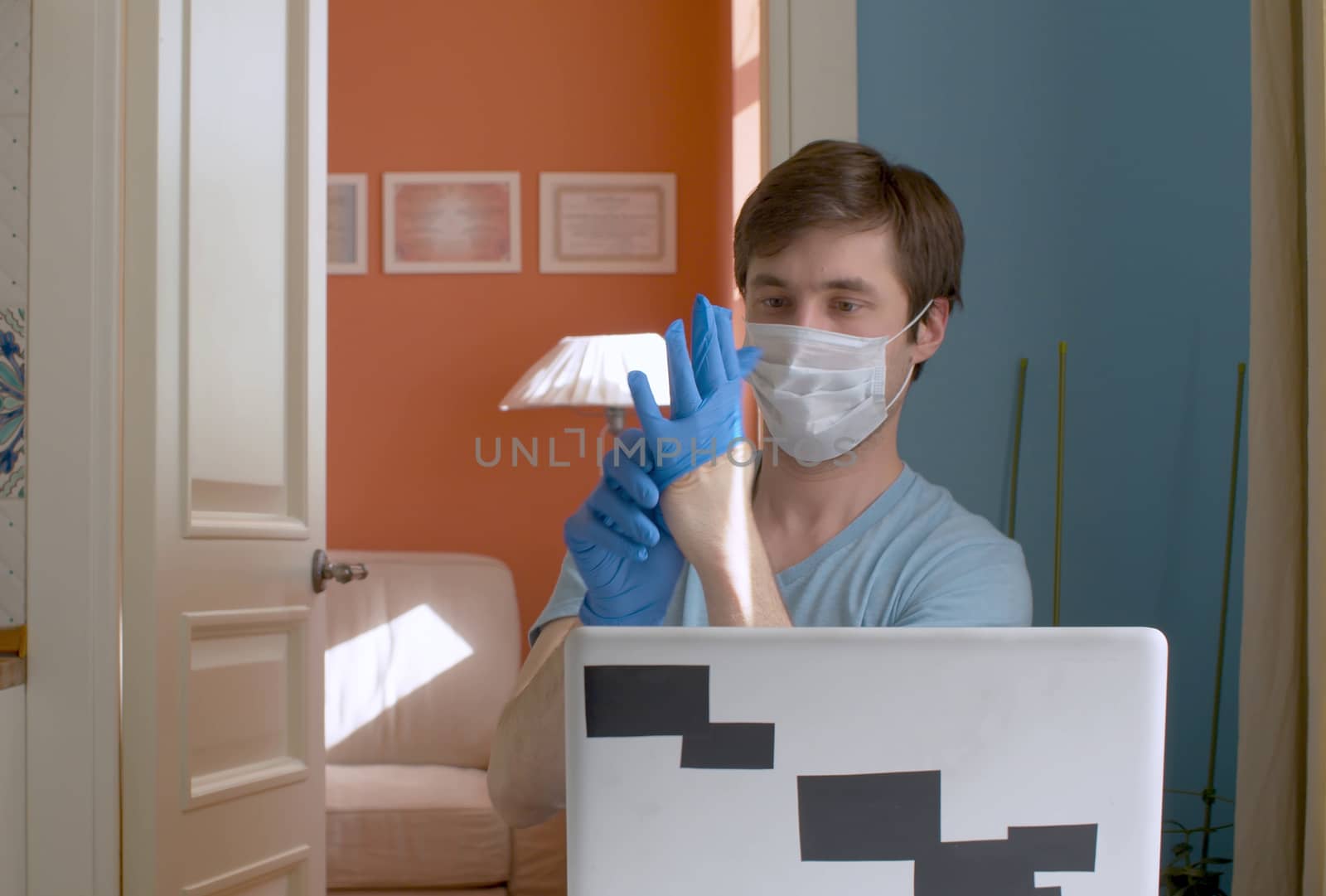 Young man working from home. He is sitting near computer and putting on blue rubber gloves. Quarantine, coronavirus epidemic.