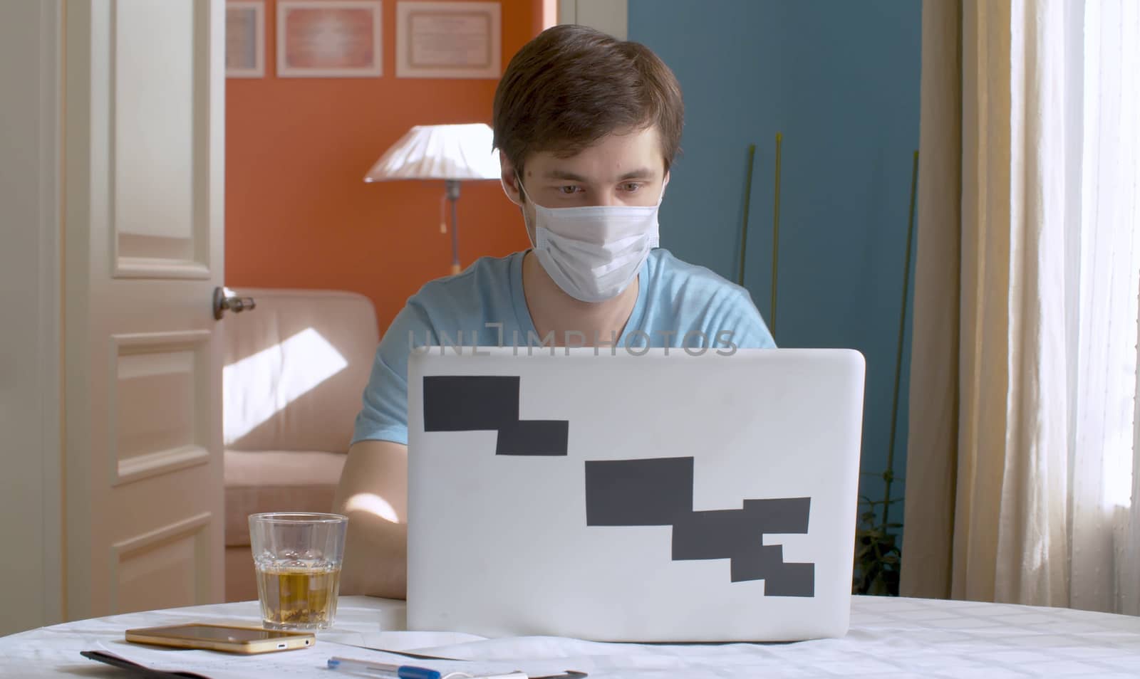 Young man working from home. He is typing on a computer in bright kitchen. Remote work during quarantine, coronavirus epidemic.