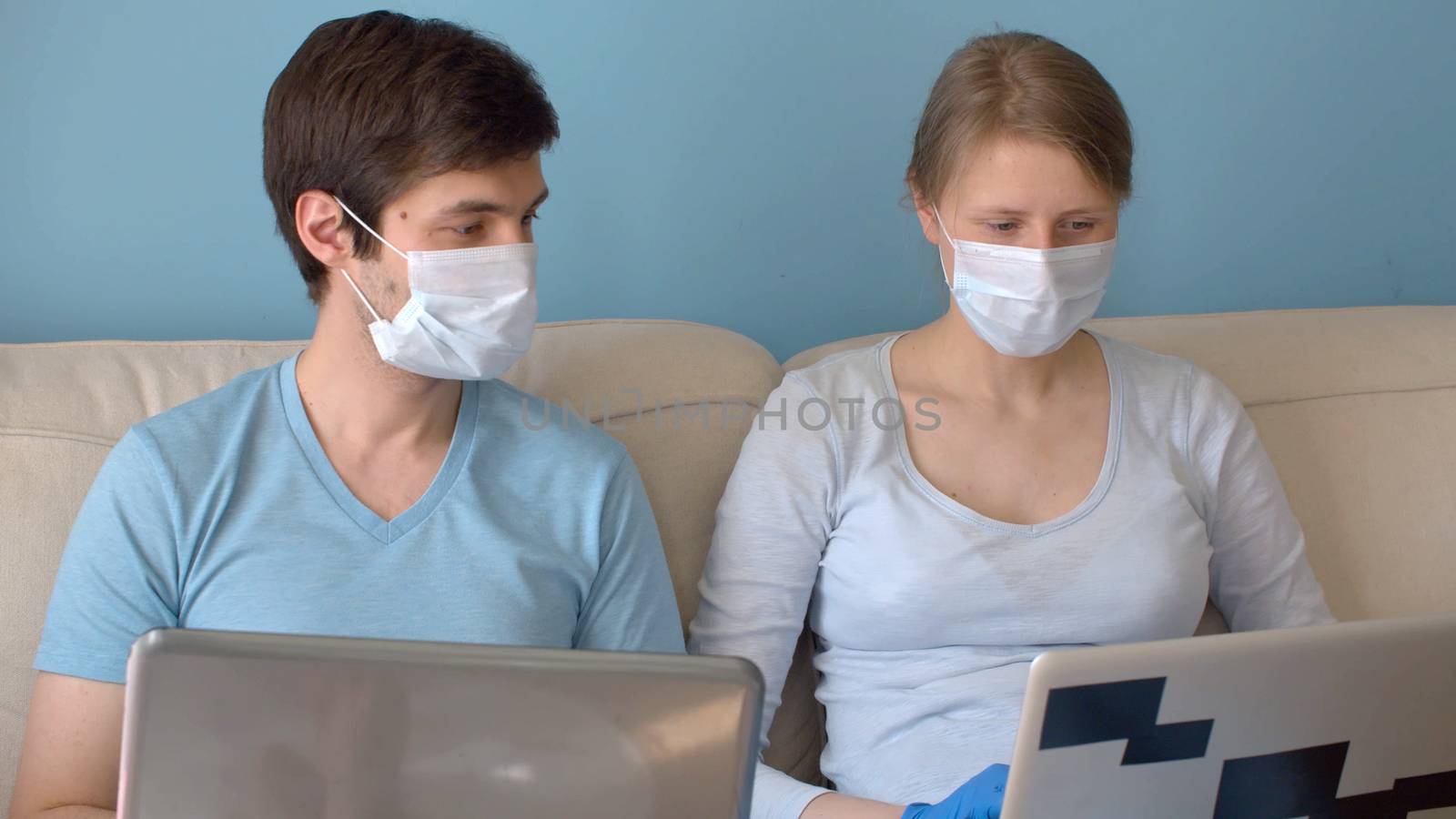 Home office. Couple on remote job. Man and woman in protective masks sitting on the sofa in bright room and typing in their computers. Quarantine, coronavirus epidemic.