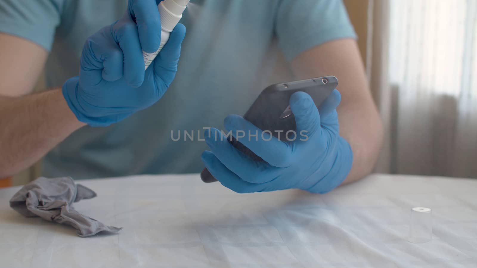 Close up male hands in protective gloves sanitaizing smart phone. He is spraying sanitizer on it. Hygiene during coronavirus epidemic. Concept of safety life.