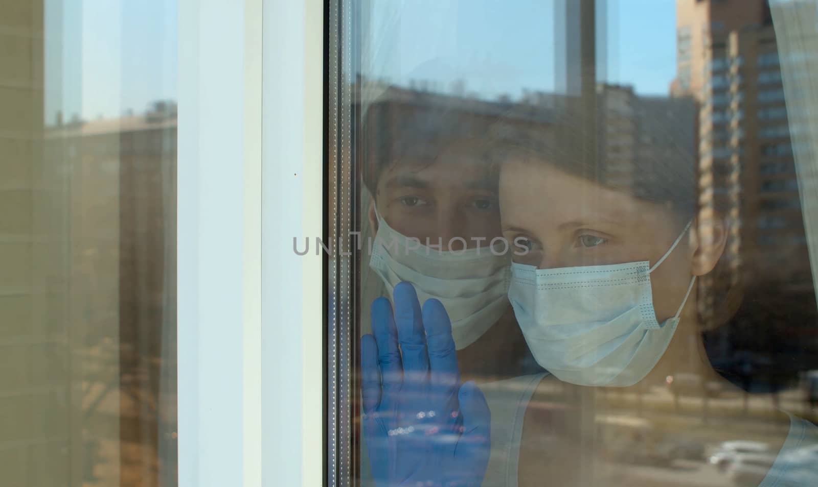 Young family portrait through the window. They are in protective masks and gloves during coronavirus quarantine. Traffic and buildings are reflected in the window glass. Covid-19 epidemic