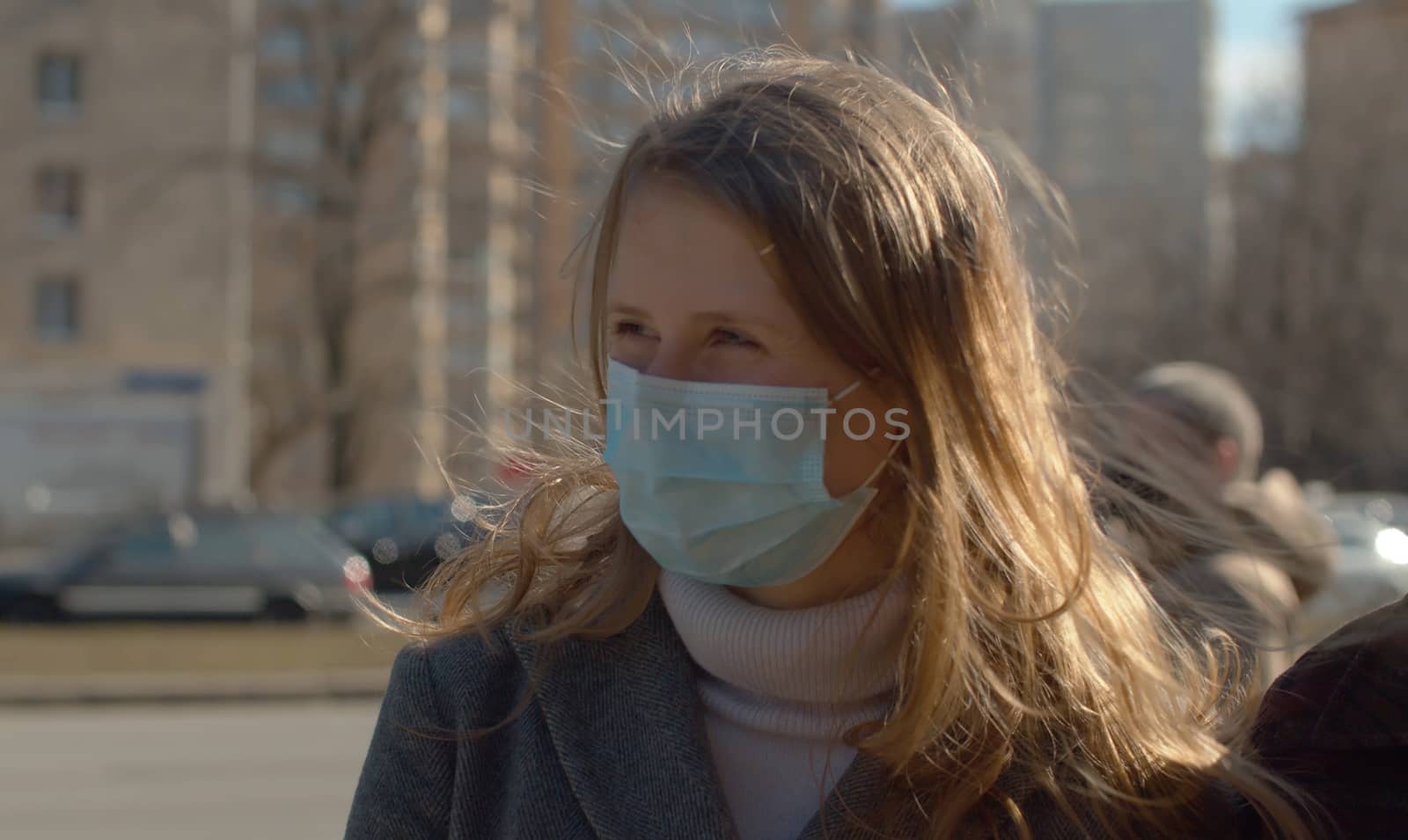 Close up portrait of a young handsome woman in protective mask outdoors on the blurry city background. The sun shines beautifully on her hair and the wind blowing them. Coronavirus epidemic