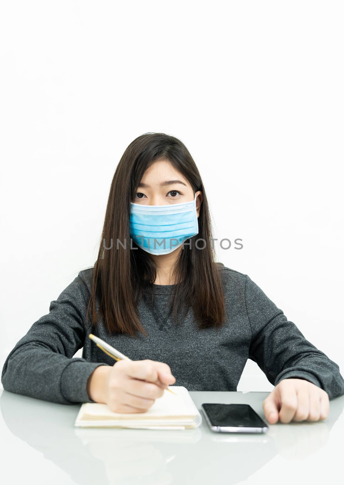 Teenage female student preparing for learnning at home sitting at a desk in front of smartphone with notepad and wear protective medical mask for protect Covid-19 or corona virus disease