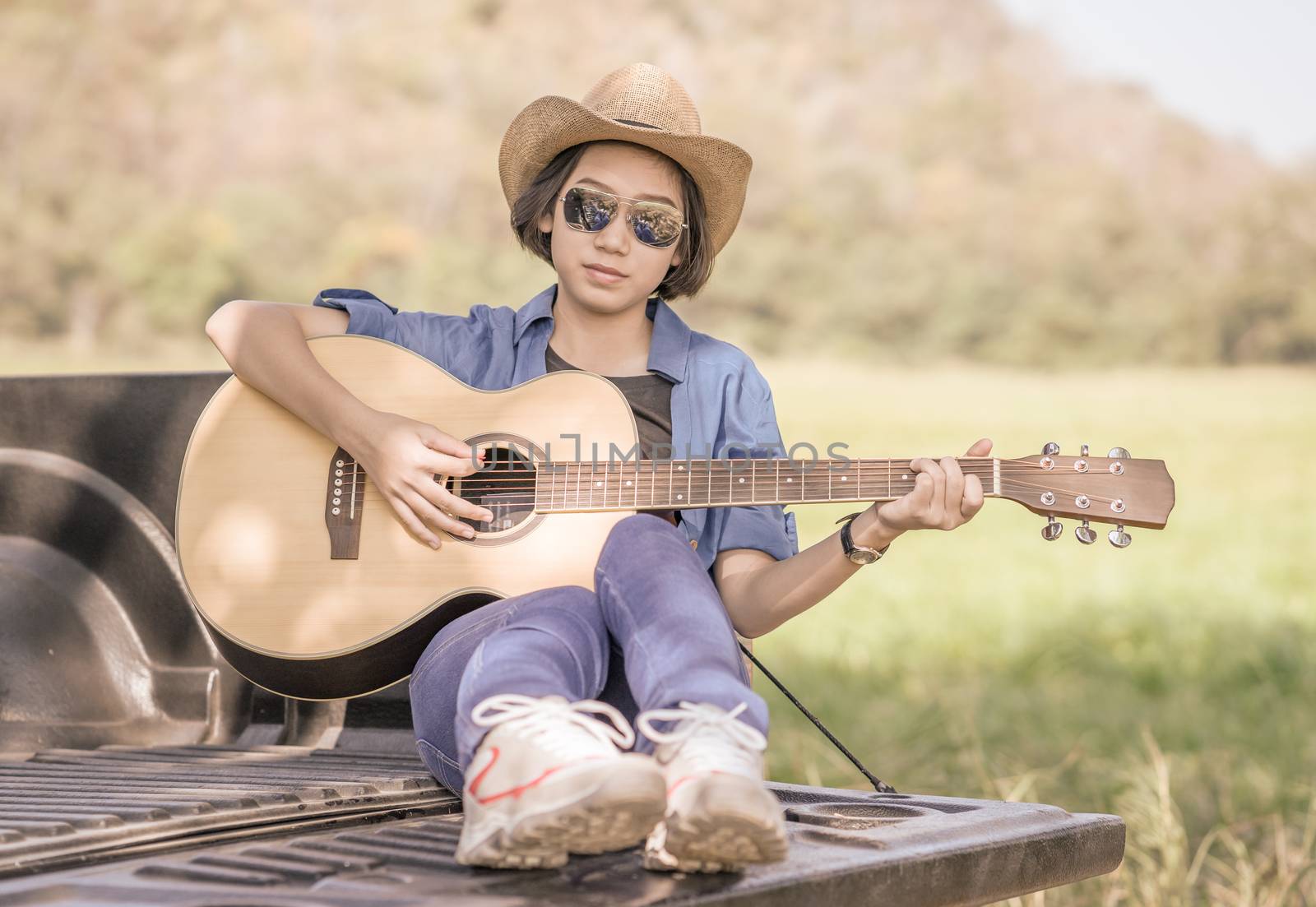 Young asian women short hair wear hat and sunglasses playing guitar ,sit on pickup truck in countryside Thailand