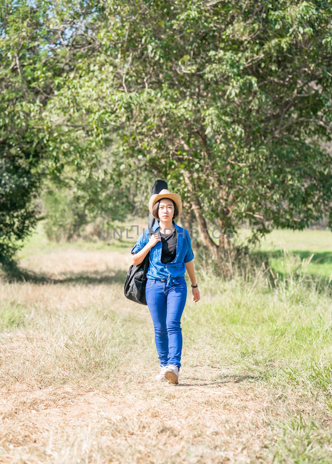 Young asian woman short hair wear hat walking and carry her guitar bag along in countryside Thailand