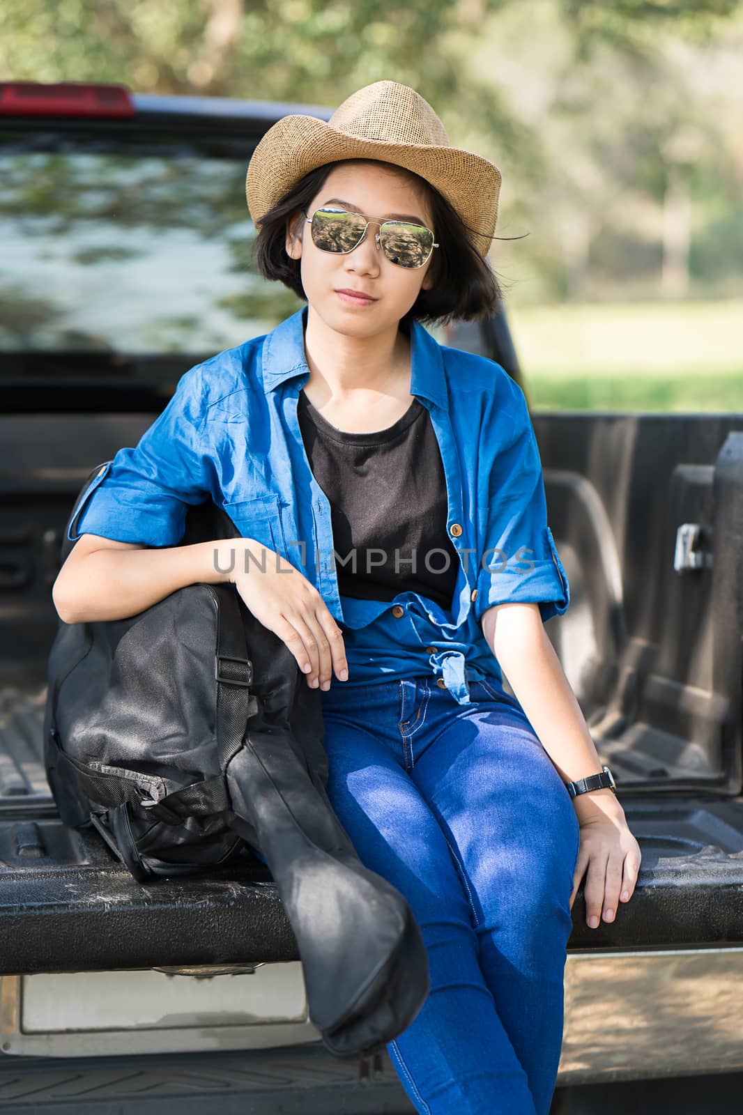 Young asian women short hair wear hat and sunglasses carry her guitar bag ,sit on pickup truck in countryside Thailand