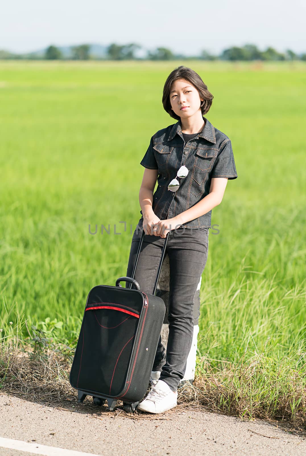 Young asian woman short hair and wearing sunglasses with luggage hitchhiking along a road in countryside Thailand