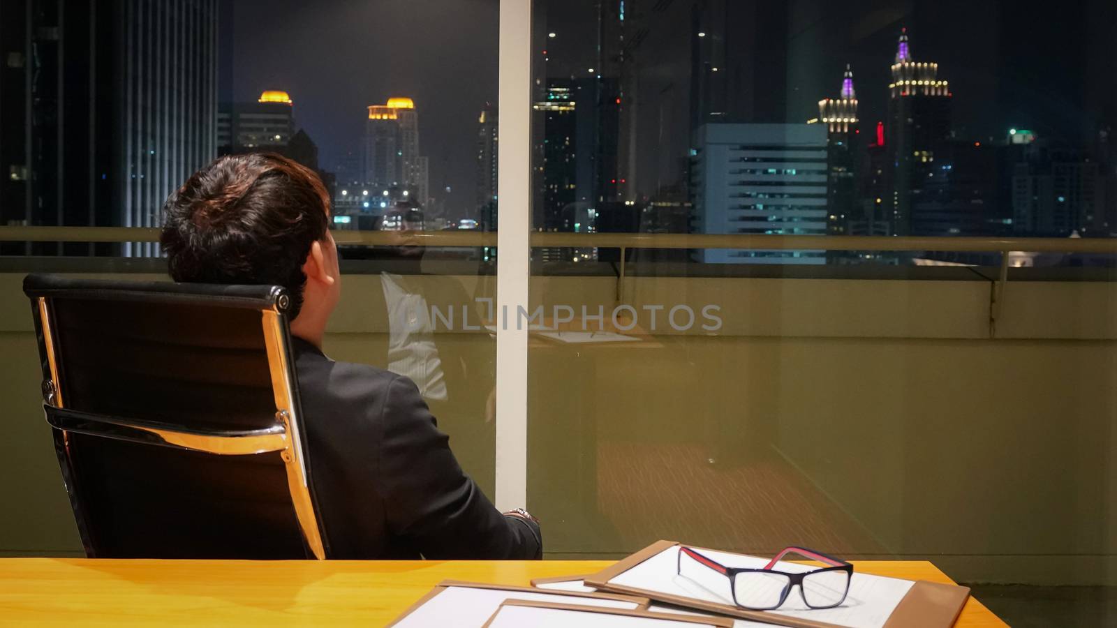 Asian businessman looking outside the window while review brief document after work. Meeting room on high floor at the night time. Bangkok's bustling downtown area.