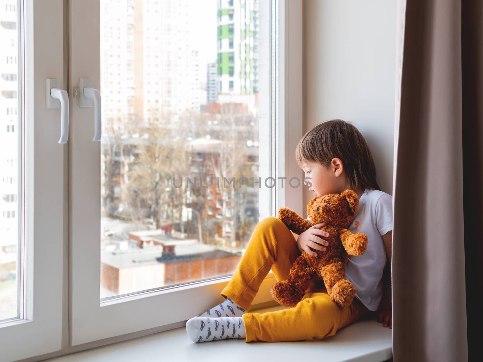Toddler boy sits with teddy bear. Kid with plush toy look through window outside. Koala bear and child on home quarantine because of coronavirus COVID-19.