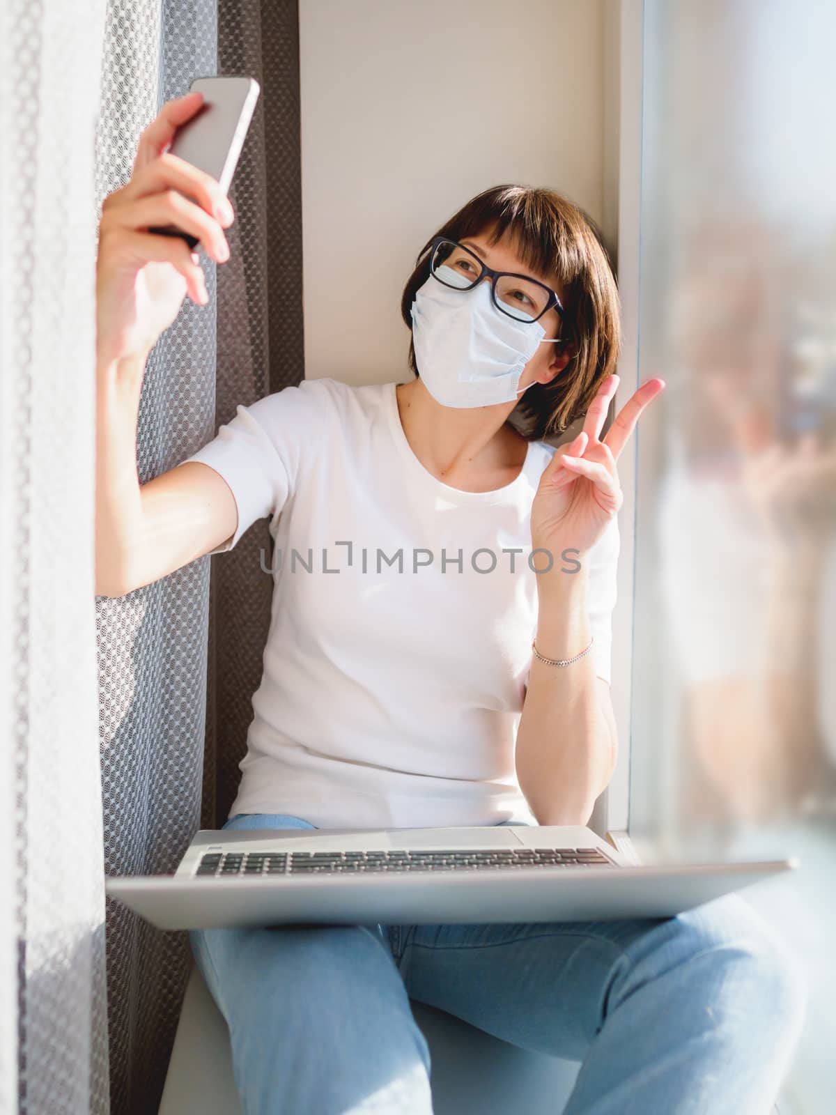 Pretty woman in medical mask works remotely from home. She is making selfie with Victory gesture on window sill with laptop on knees. Lockdown quarantine because of coronavirus COVID19. Self isolation at home.