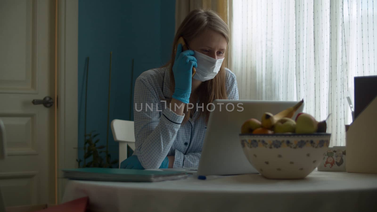 Portrait of a young woman working from home. Lady in a protective mask and gloves looking in a computer and talking by smart phone. Remote job, quarantine. COVID-19 pandemic