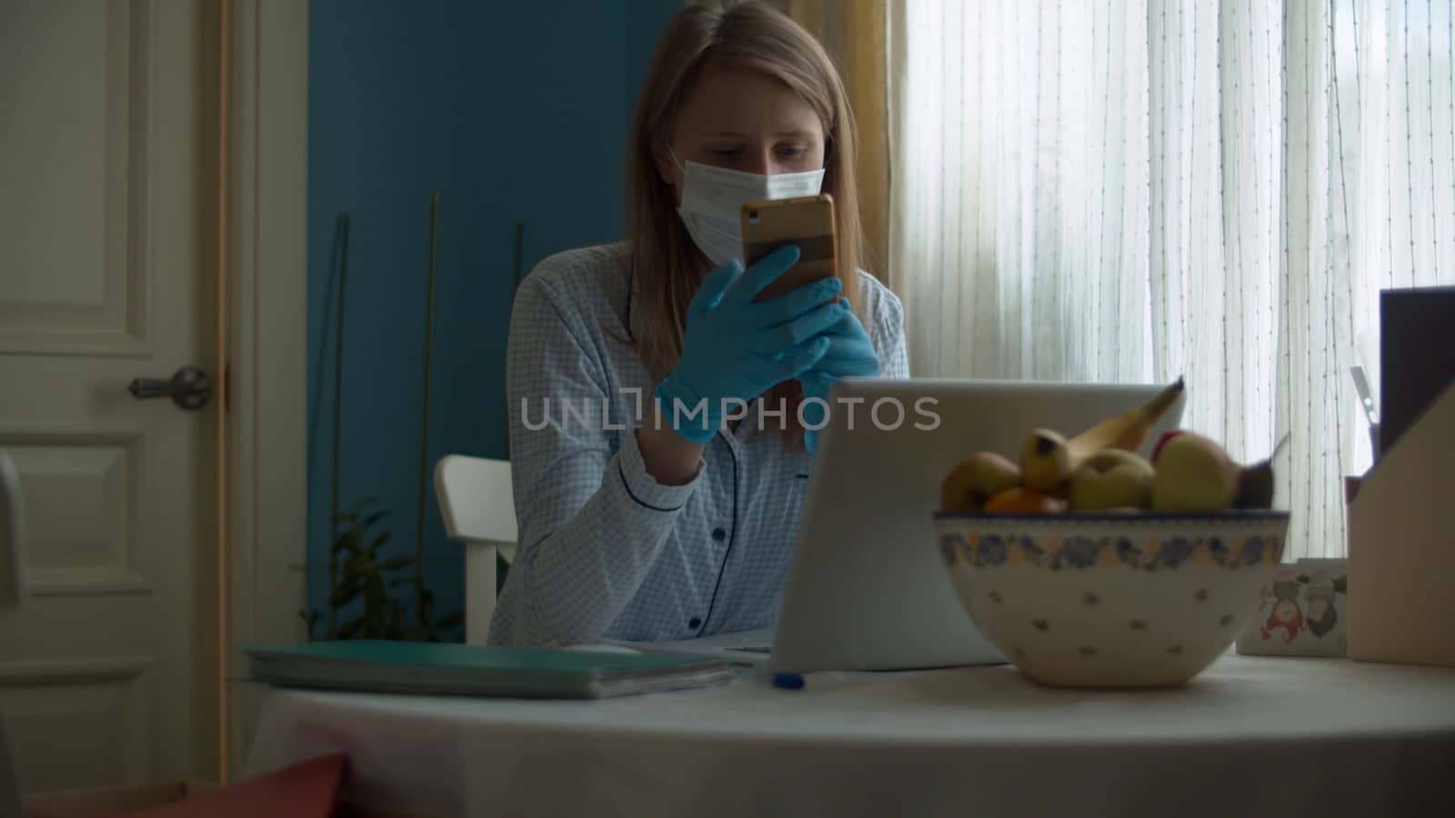 Portrait of a young woman working from home. Lady in a protective mask and gloves looking in a computer and talking by smart phone. Remote job, quarantine. COVID-19 pandemic