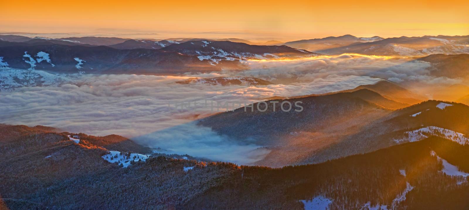 Winter sunrise panorama from the summit in Romanian Carpathians; misty valley and sun rays.