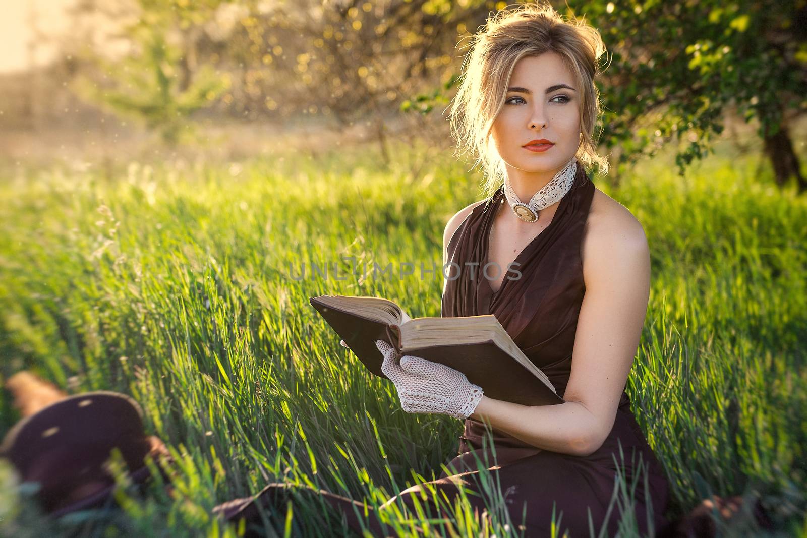 young blonde girl in a brown vintage dress and top hat reads a book in spring garden