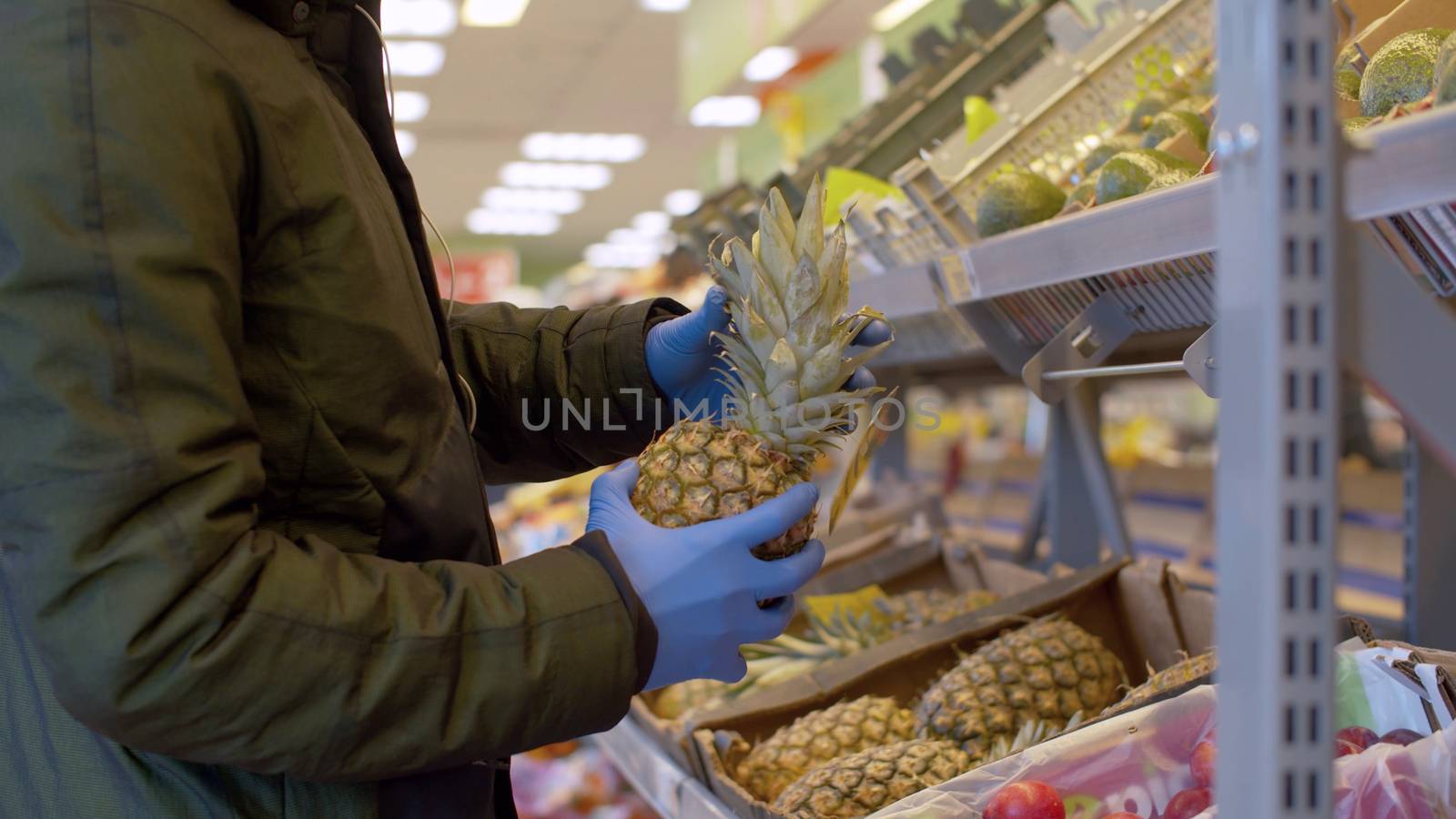 A man in a protective mask and gloves choosing pineapple in the supermarket. Coronavirus epidemic in the city. COVID-19 pandemic