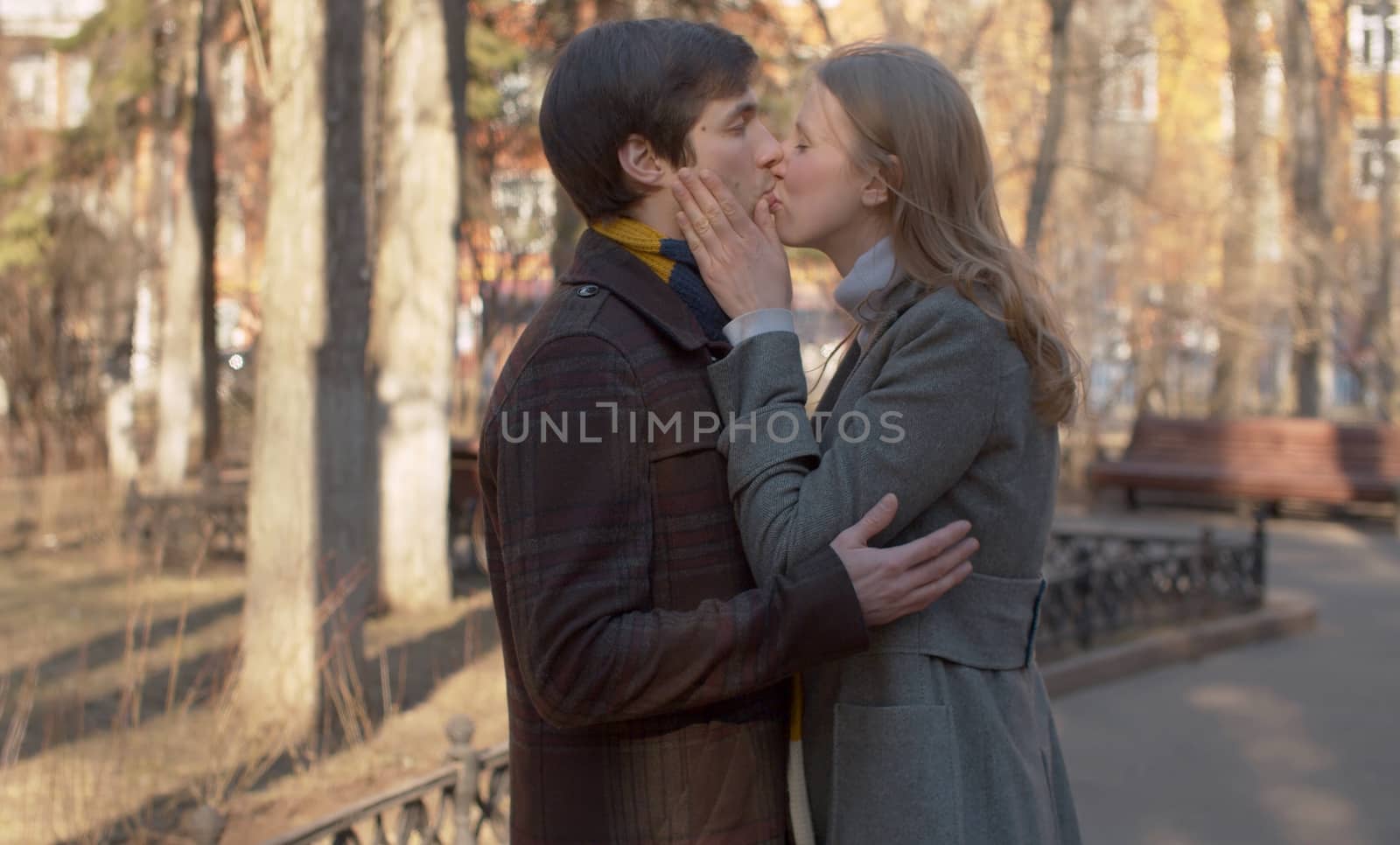 Beautiful couple in love kissing in the park, young blond girl in a gray coat and man in brown coat and colorful scarf. Spring sunny day.