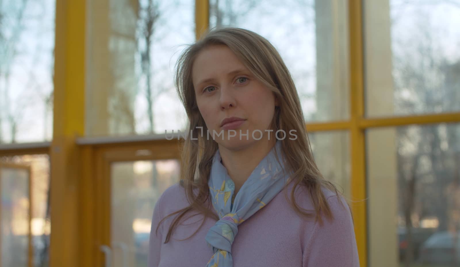 Close up portrait of a young attractive serious woman standing against modern glass building. Blond woman with long hair outdoors