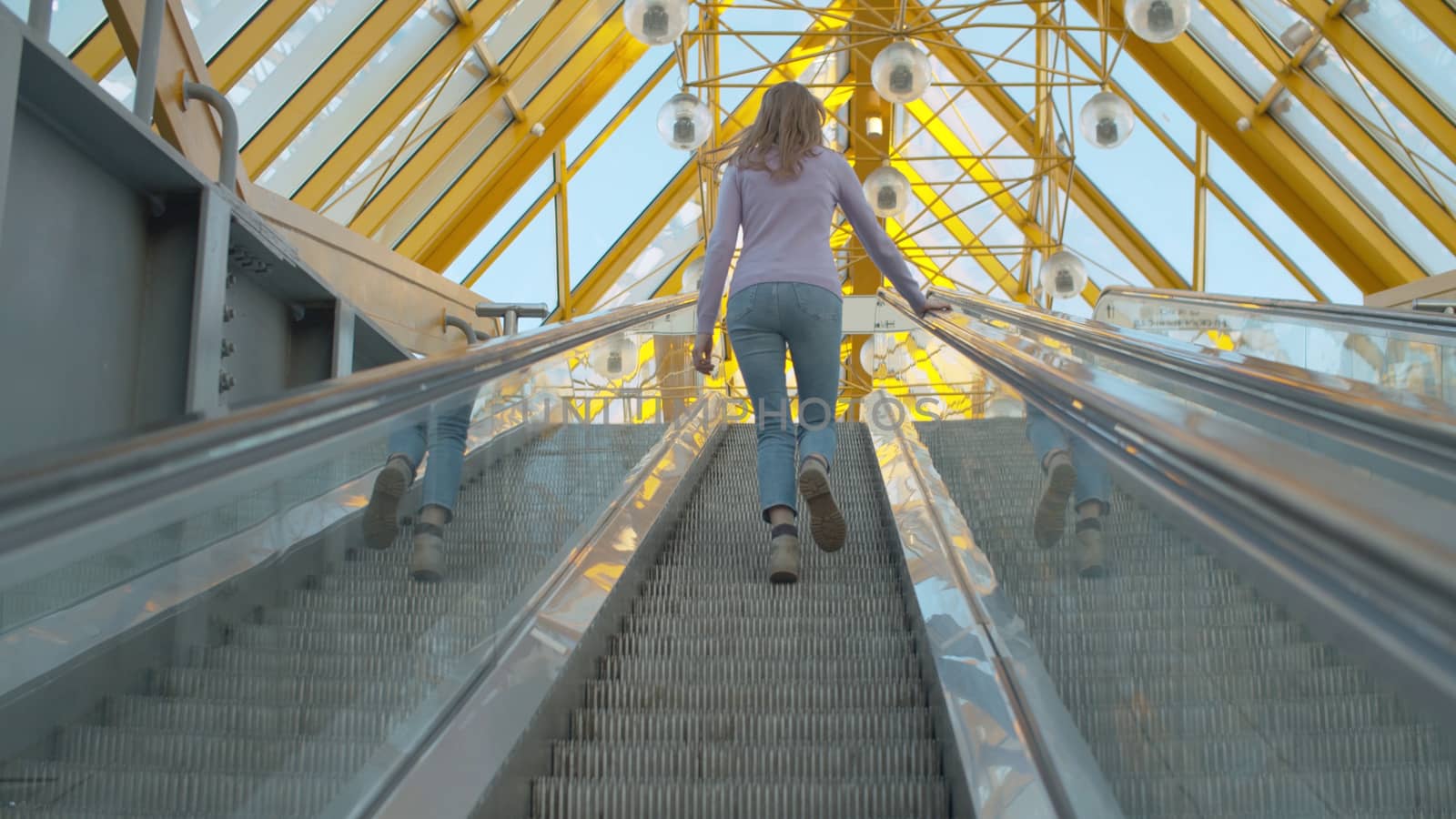 A young woman climbing up the moving escalator. Rear view, follow shot. Blond woman with long hair inside modern building