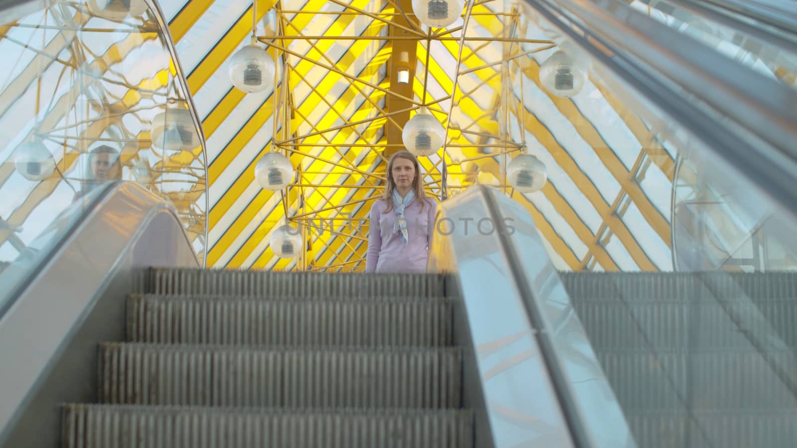 A young woman standing on the top of the moving escalator. Camera moving to she. Blond woman with long hair inside modern building