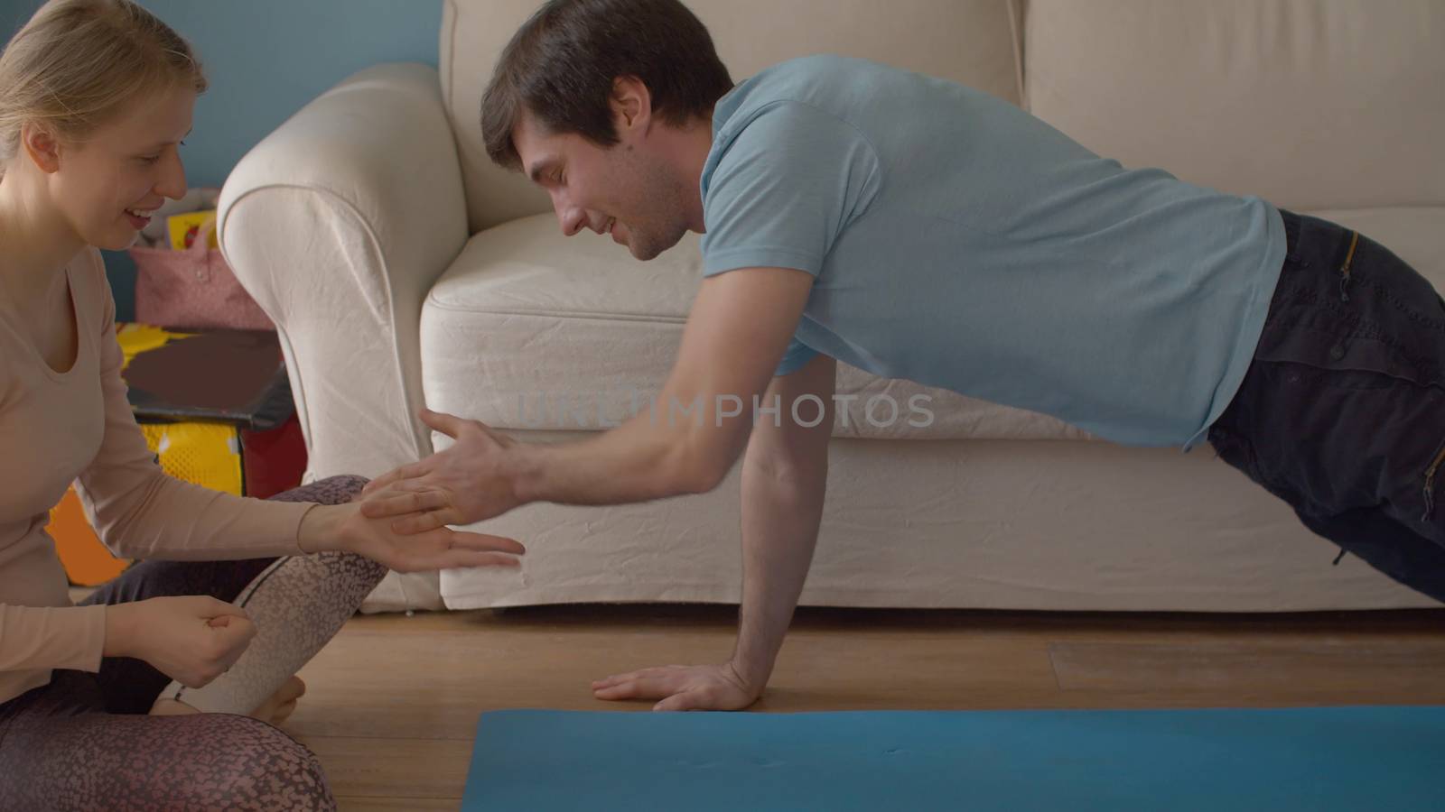 A young cheerful family spends time at home. The husband does push-ups with clapping, the wife sitting nearby. Both laughing. Coronavirus epidemic