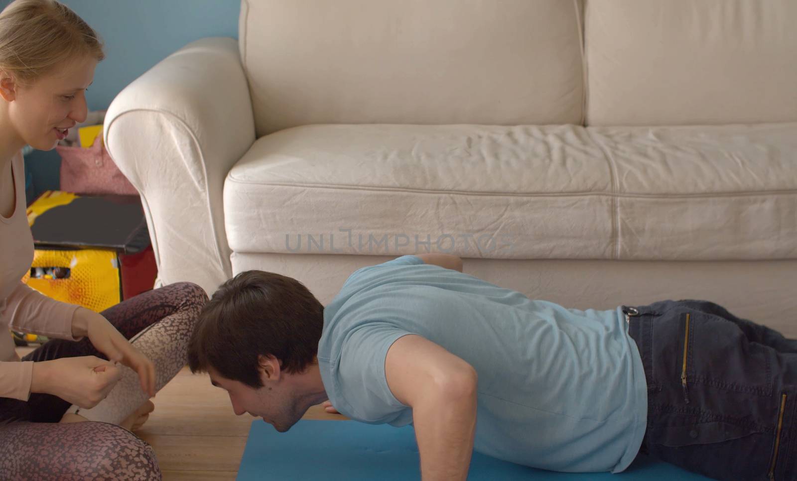 A young cheerful family spends time at home. The husband does push-ups with clapping, the wife sitting nearby. Both laughing. Coronavirus epidemic