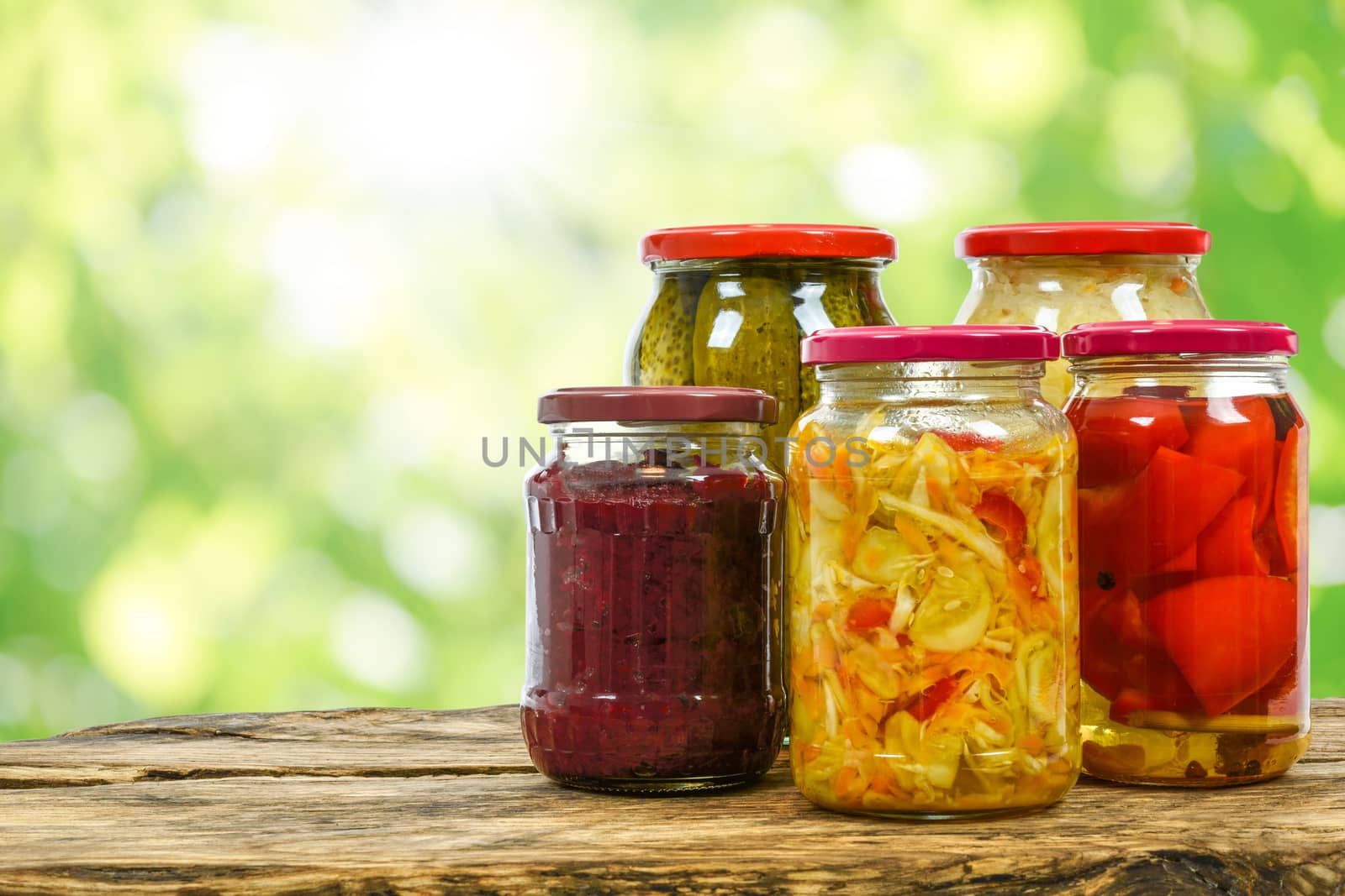 Composite image of jars with variety of homemade pickled vegetables on a wooden vintage table and on the blurred green garden background.