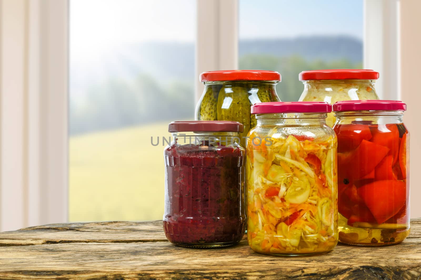 Composite image of jars with variety of homemade pickled vegetables on a wooden vintage table and on the background of a blurred window in the kitchen.