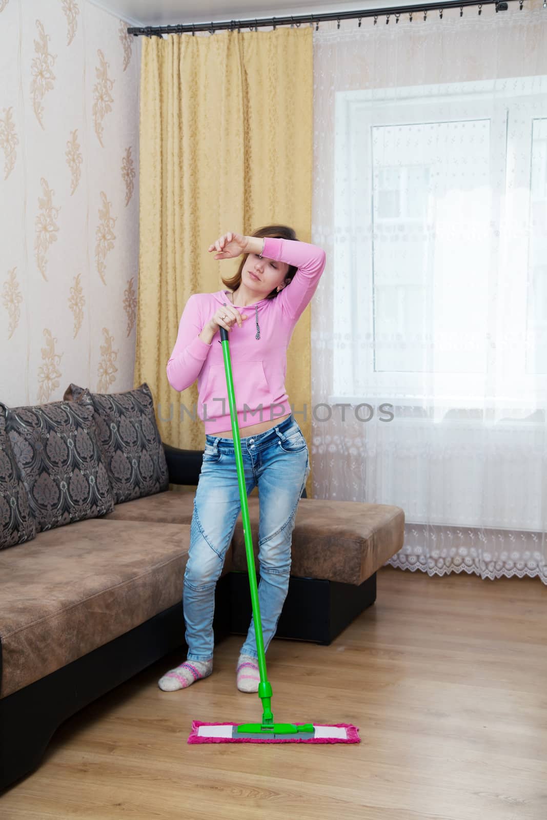 young tired woman in a pink blouse and blue jeans washing wooden floor with a mop