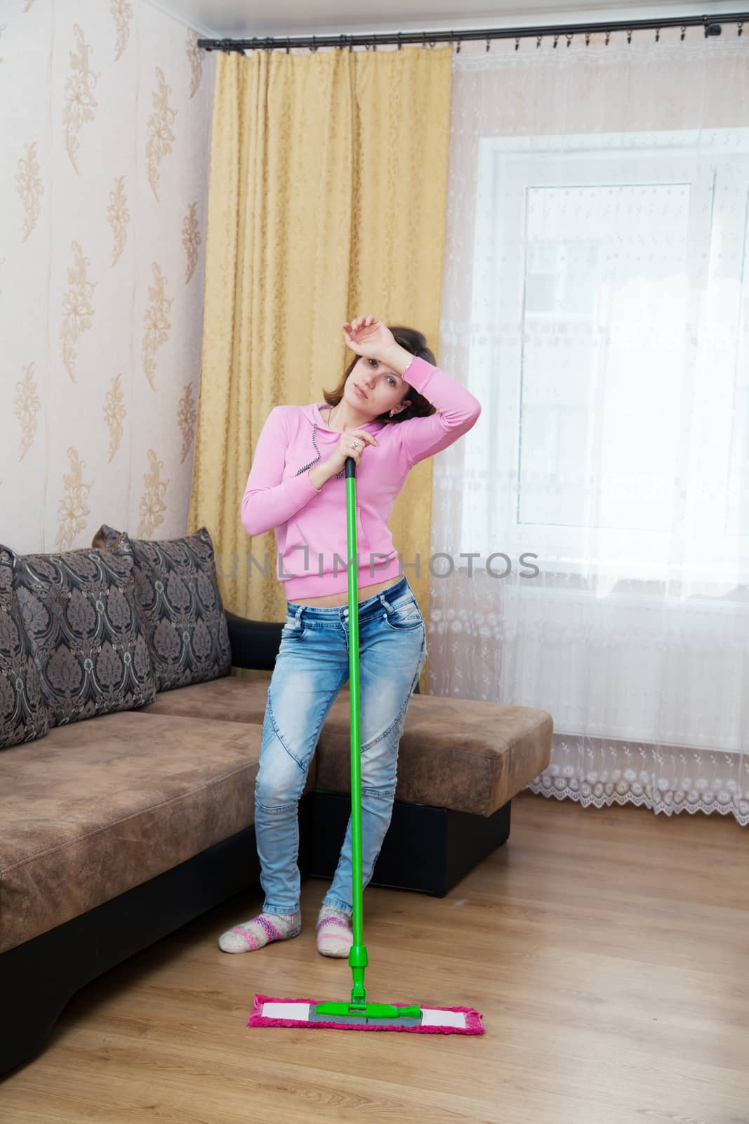 young tired woman in a pink blouse and blue jeans washing wooden floor with a mop