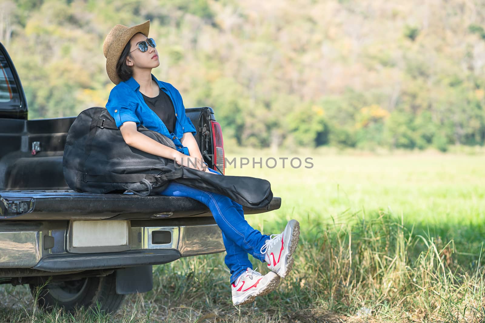 Young asian women short hair wear hat and sunglasses carry her guitar bag ,sit on pickup truck in countryside Thailand