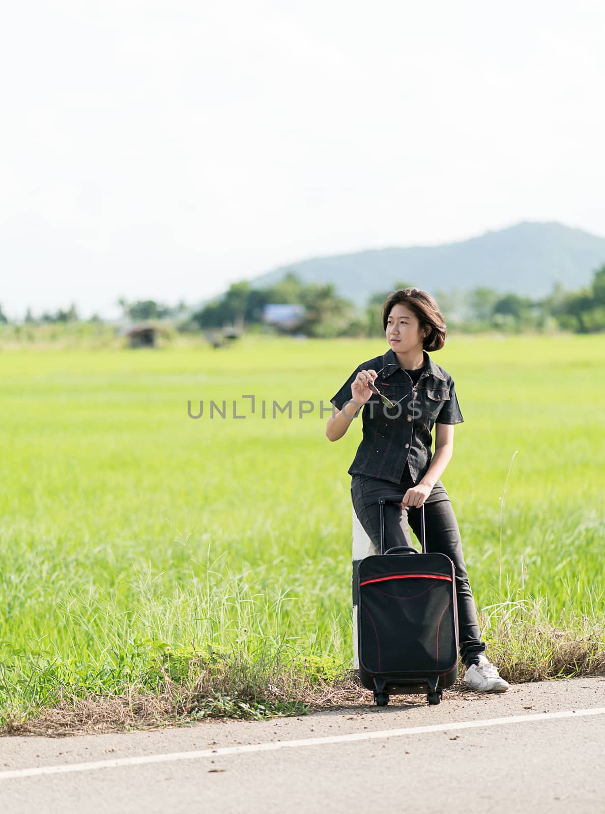 Young asian woman short hair and wearing sunglasses with luggage hitchhiking along a road in countryside Thailand