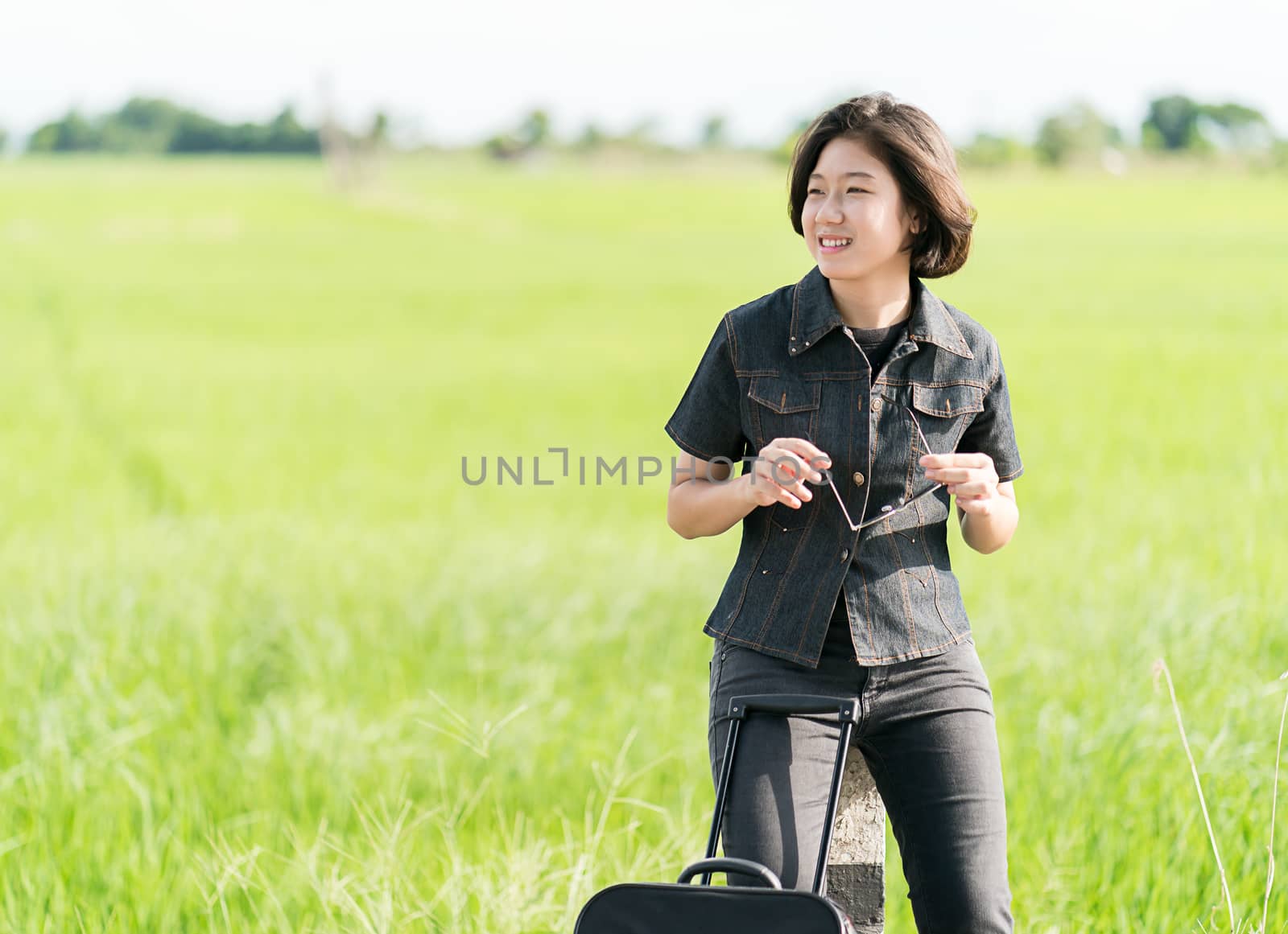 Young asian woman short hair and wearing sunglasses with luggage hitchhiking along a road in countryside Thailand