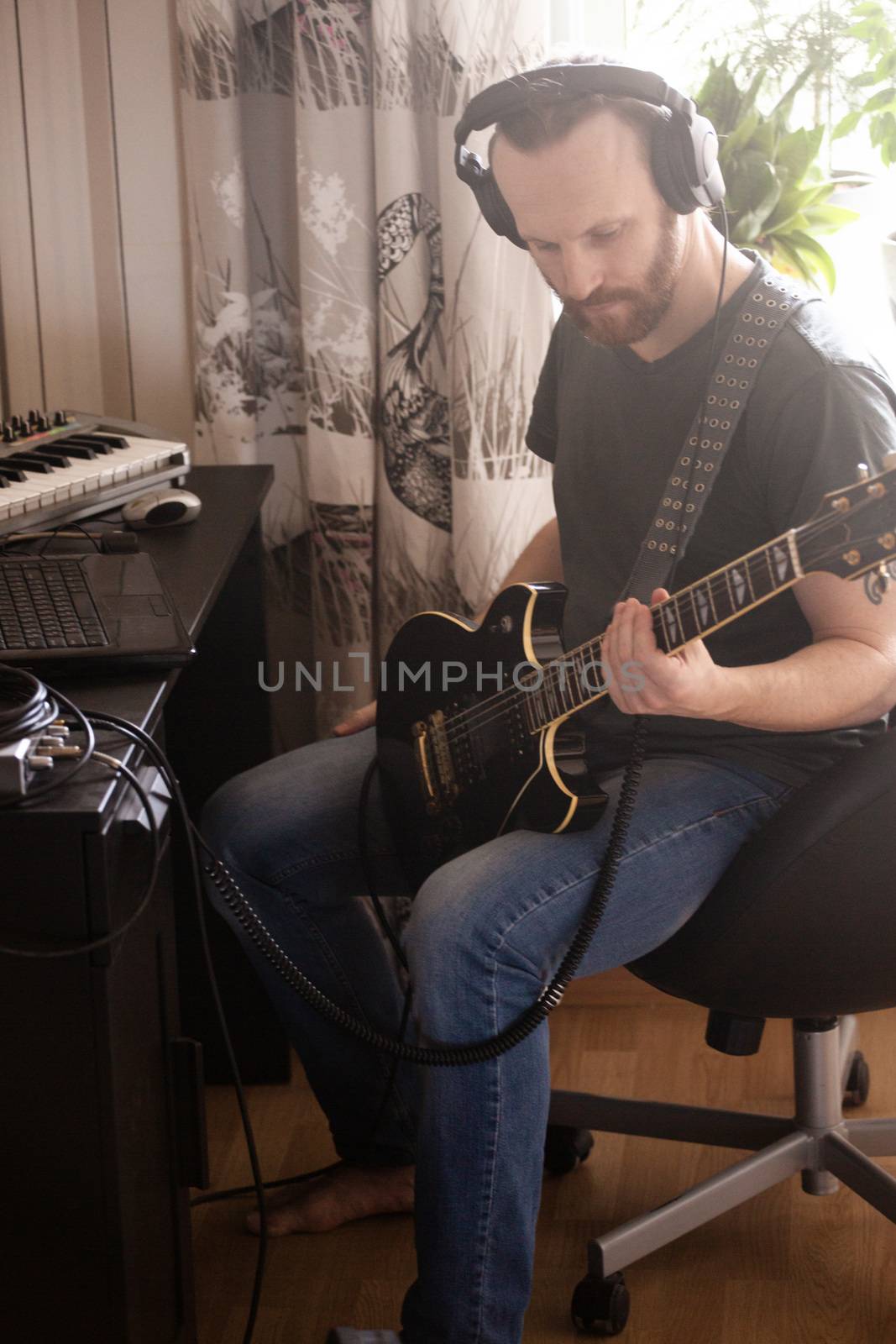 Bearded man musician playing music and composing a song with electric guitar piano and laptop computer while sitting in living room, corona virus quarantine stay home concept