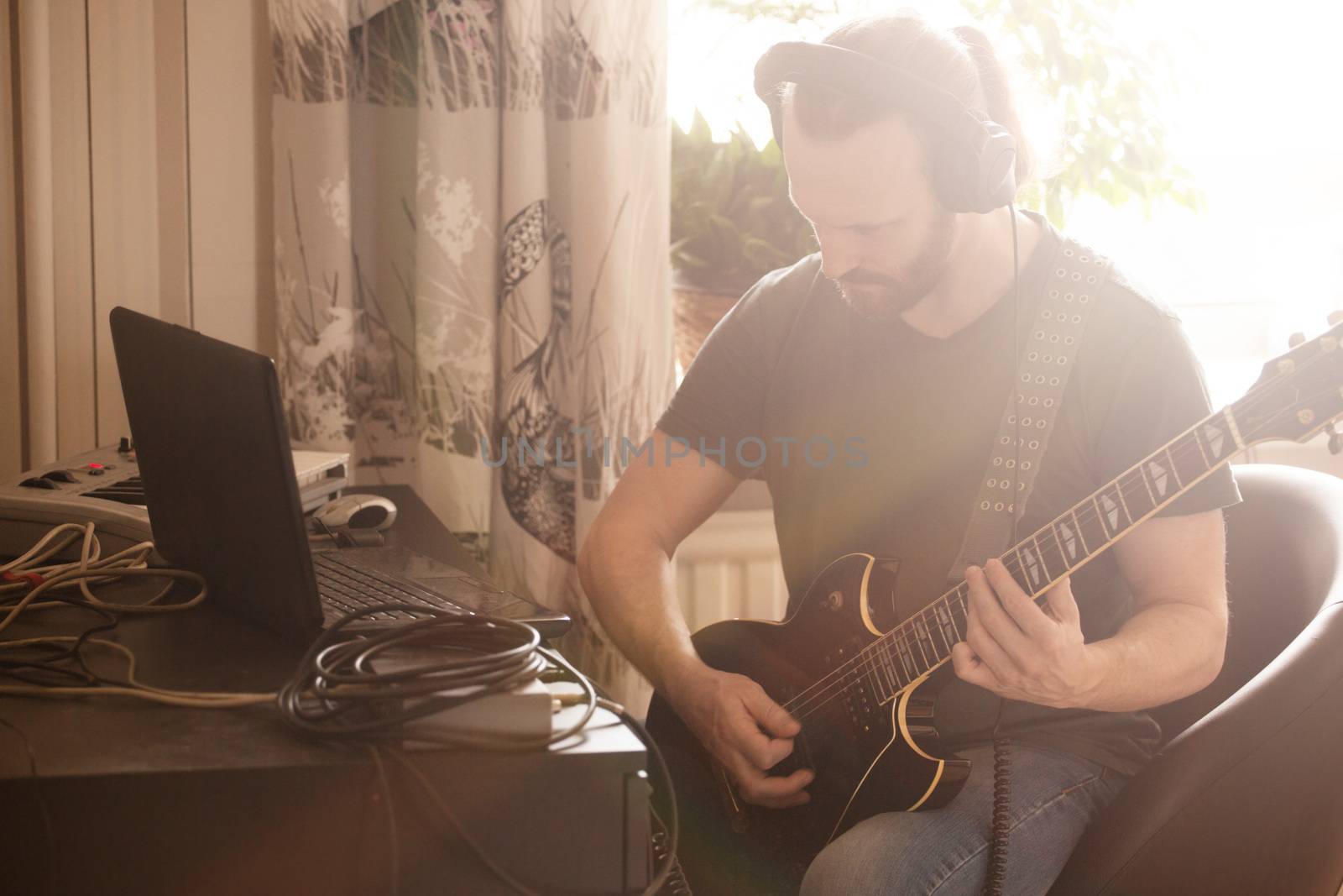 Bearded man musician playing music and composing a song with electric guitar piano and laptop computer while sitting in living room, corona virus quarantine stay home concept