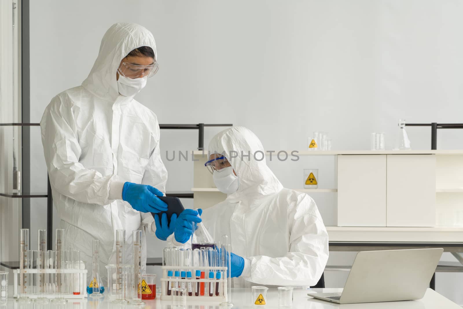 Epidemiological researchers in virus protective clothing use tablet computers to research chemical compounds on the internet. Working atmosphere in chemical laboratory.