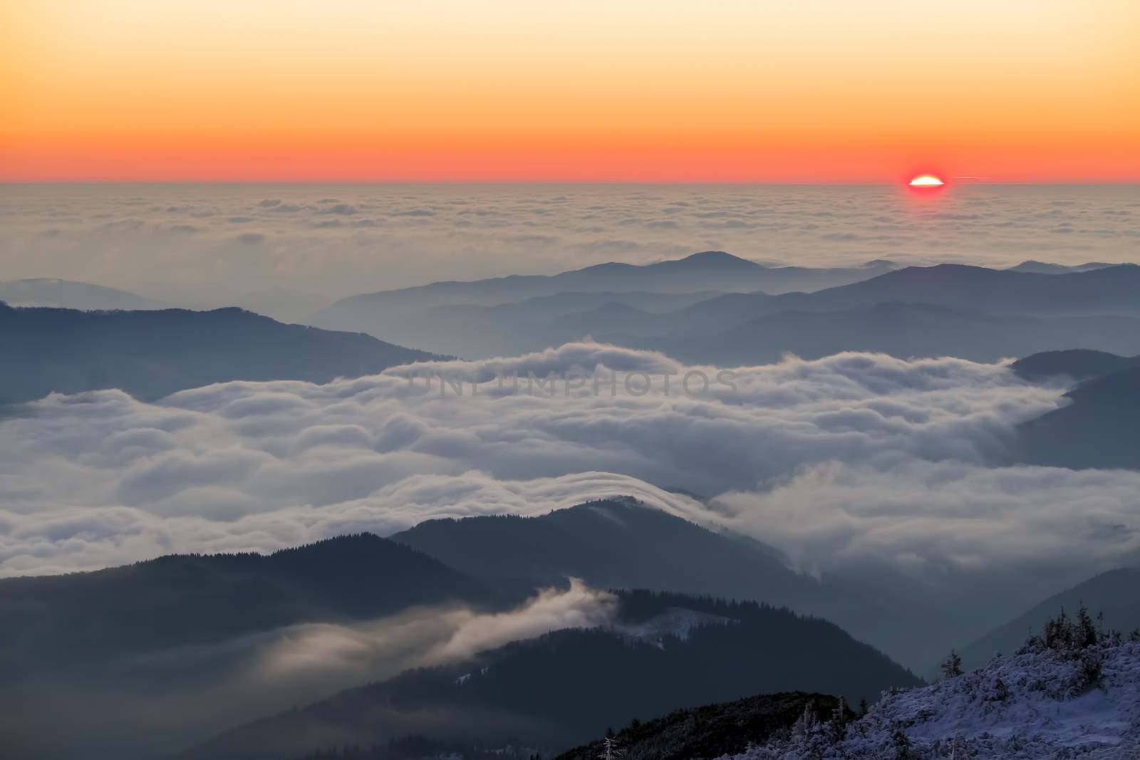 Low clouds on mountain valley and colorful sunrise viewed from mountain peak in winter, Romanian Carpathians.