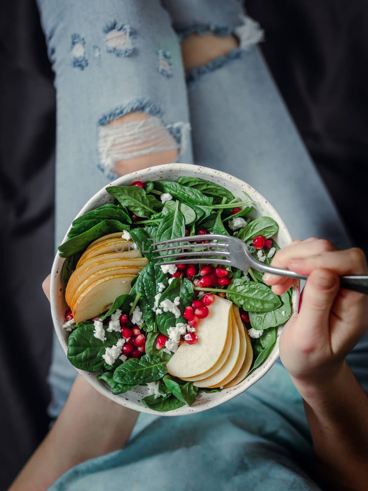 Woman in jeans holding vegan salad bowl with spinach, pear, pomegranate, cheese. Vegan breakfast, vegetarian food, diet concept. Girl in jeans holding fork with knees and hands visible