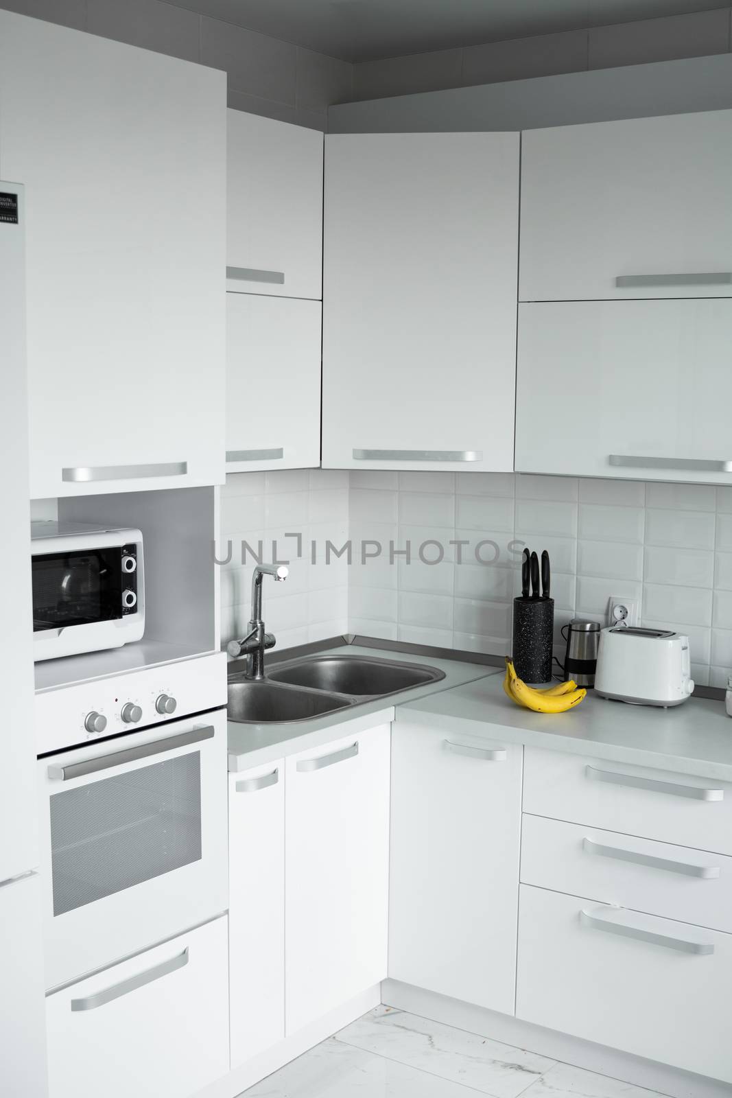 Comfortable white kitchen with a white lacquered facades. Modern kitchen clean interior design. Refrigerator, kitchen oven, microwave oven and sink. Kitchen supplies