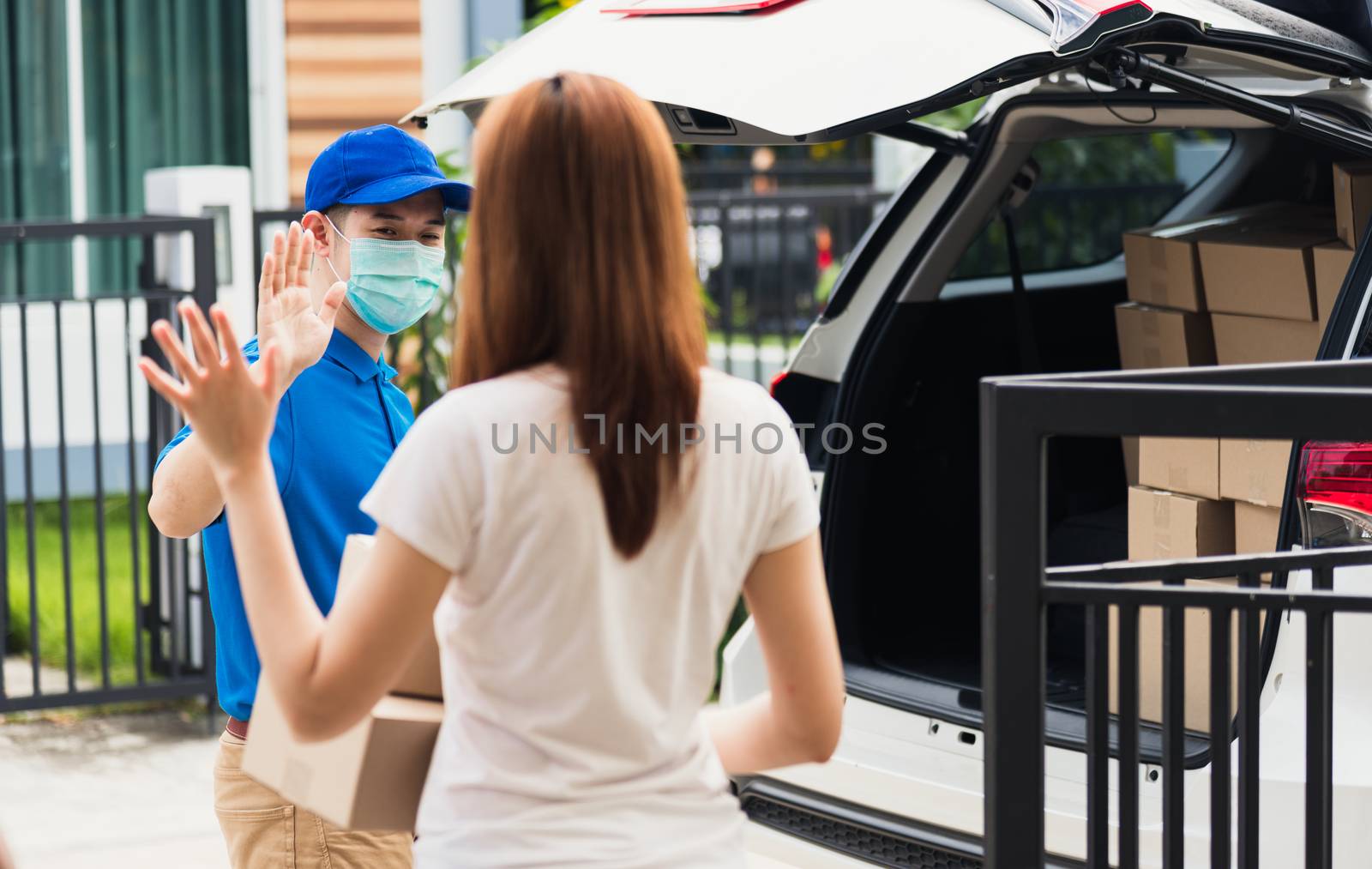 Asian young delivery man raises hand for bye after woman customer received boxes, both protective face mask under curfew quarantine pandemic coronavirus COVID-19 at front home door