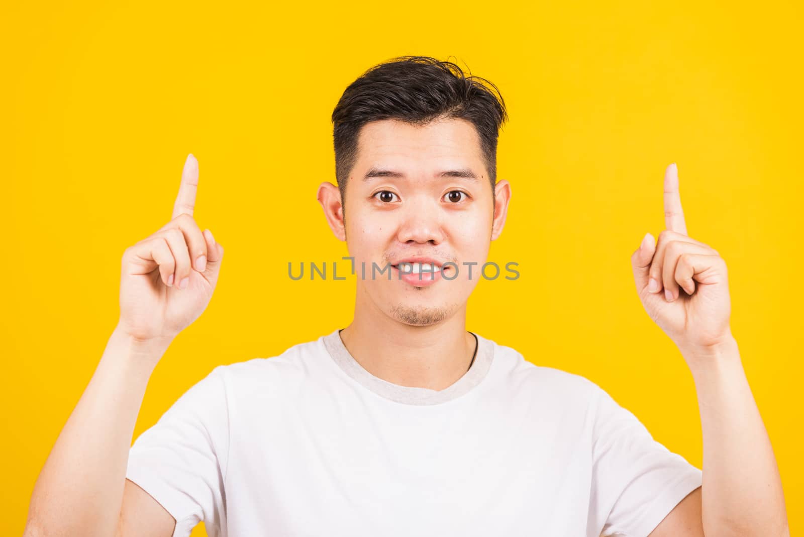 Portrait happy Asian handsome young man smiling standing wearing white t-shirt pointing finger to above UP side he looking to camera, studio shot isolated yellow background