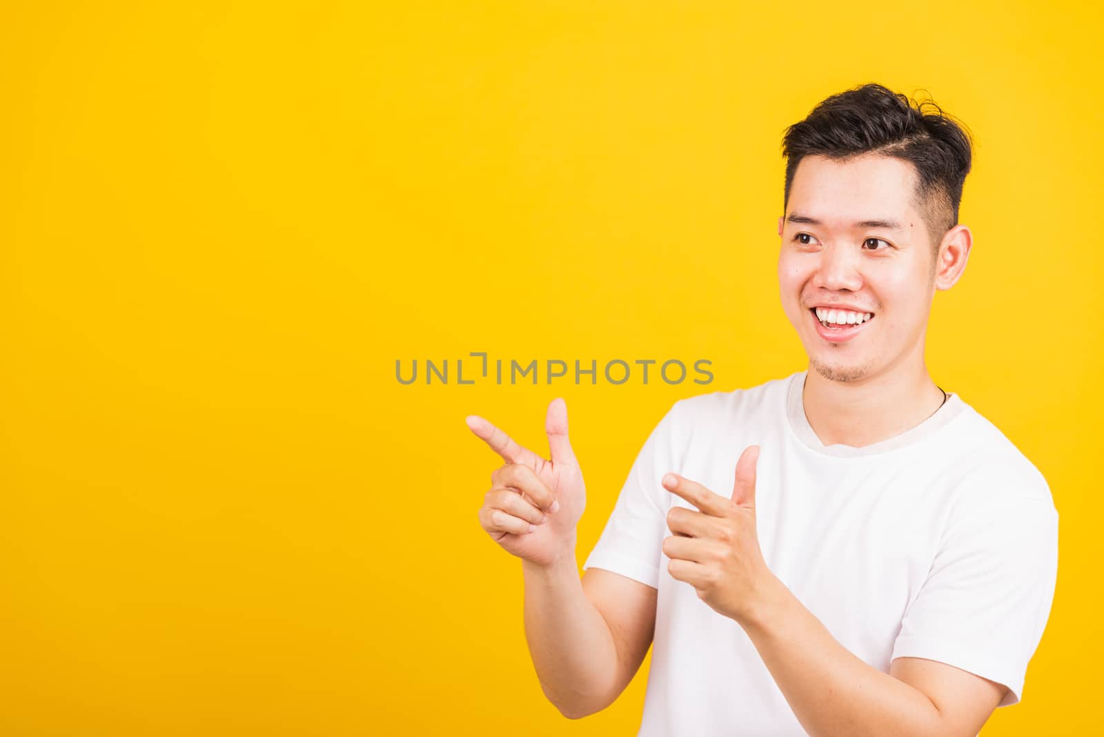 Portrait happy Asian handsome young man smiling standing wearing white t-shirt pointing finger to the side away he looking to space, studio shot isolated yellow background
