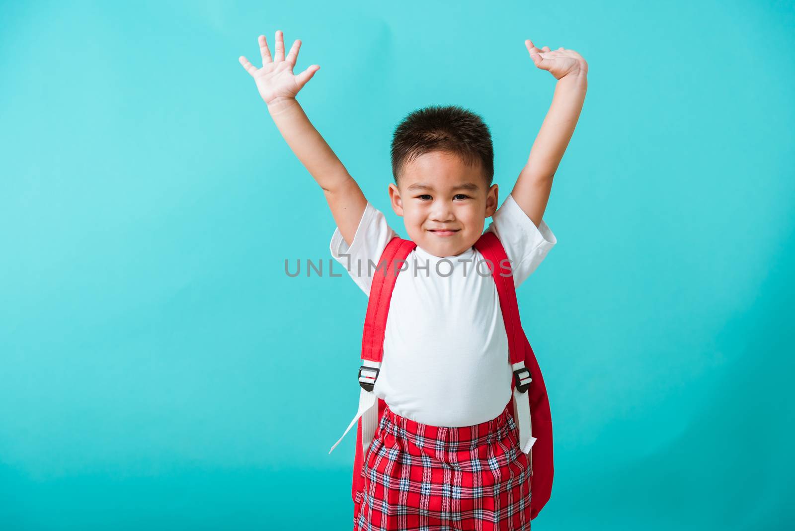 Back to school. Portrait happy Asian cute little child boy in uniform smile raise hands up glad when go back to school, isolated blue background. Kid from preschool kindergarten with school backpack