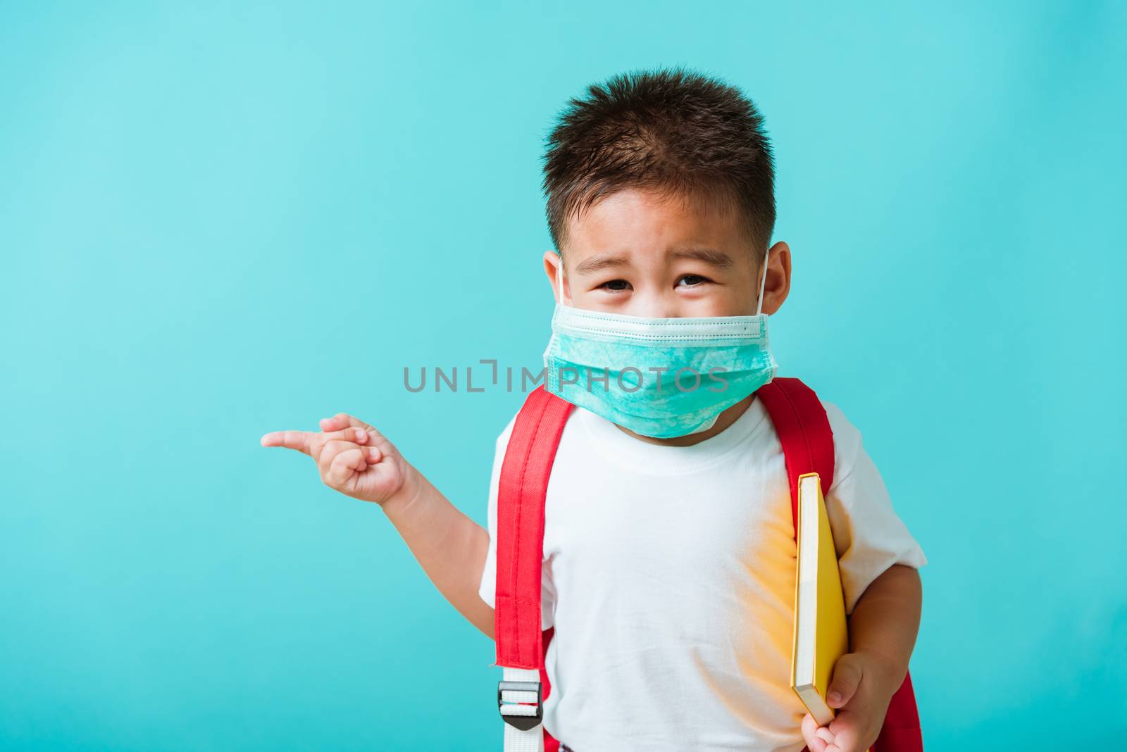 Back to school coronavirus Covid-19 education. Portrait Asian little child boy kindergarten wear face mask protective and school bag hold book before going to school pointing to side away, studio shot