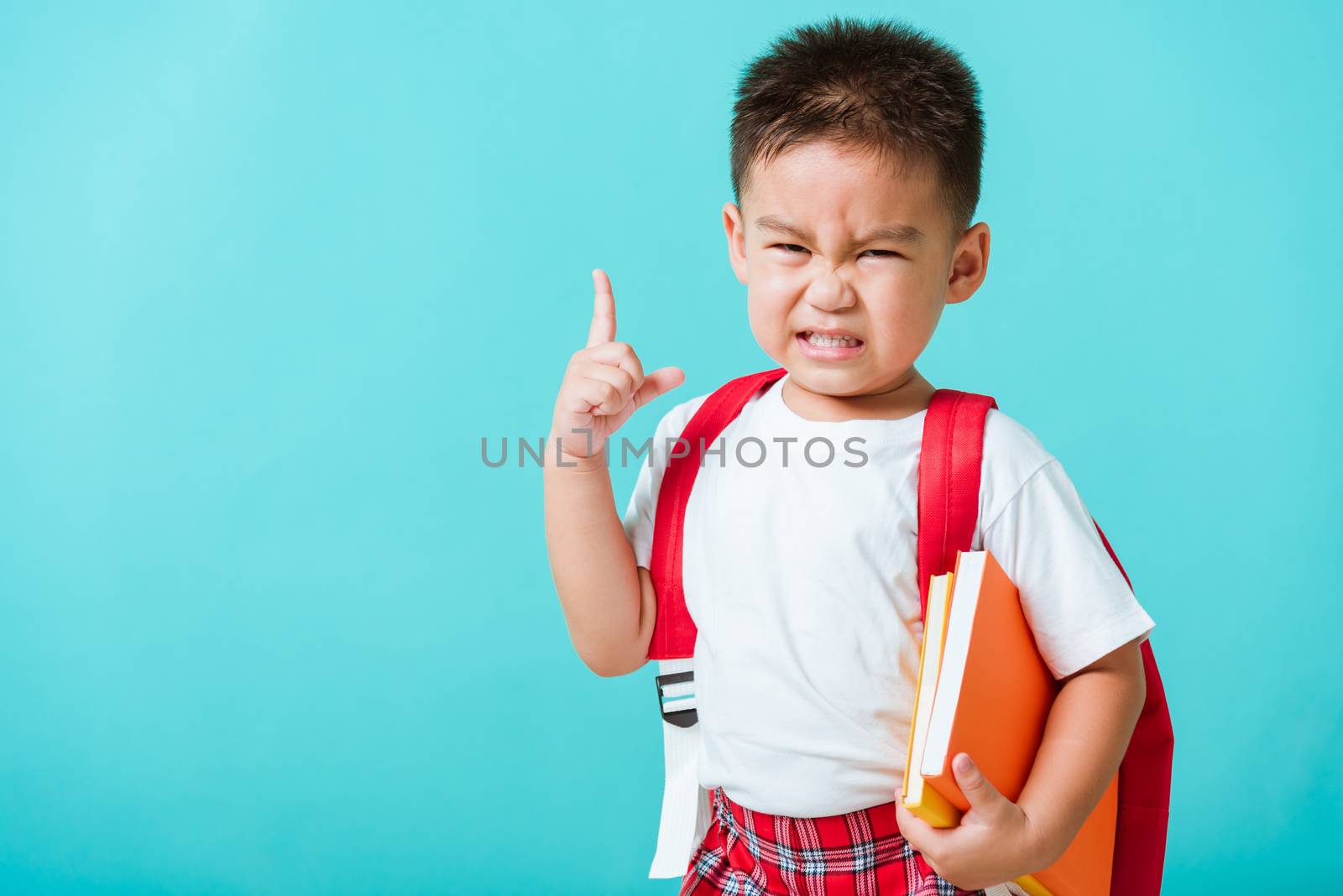 Back to school concept. Portrait Asian cute little child boy face serious hug books thinking and point finger up space, isolated blue background. Kid from preschool kindergarten with school bag