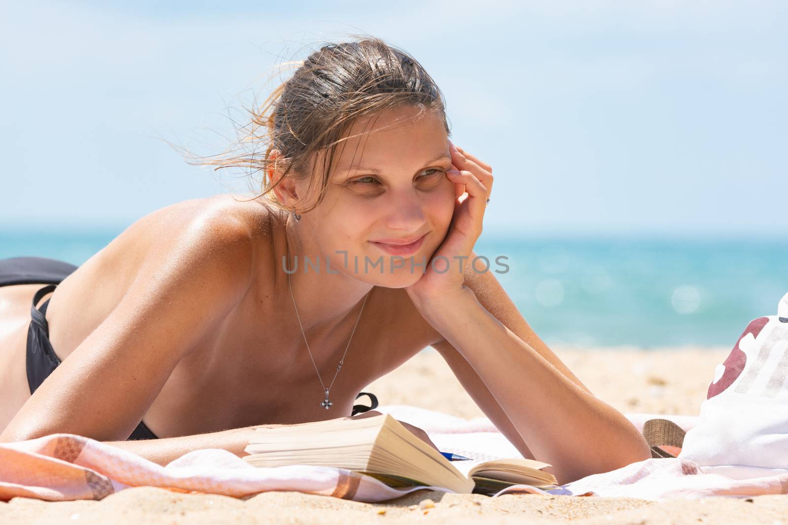 Girl dreamily looks into the distance lying on a sandy beach by the sea with a book by Madhourse
