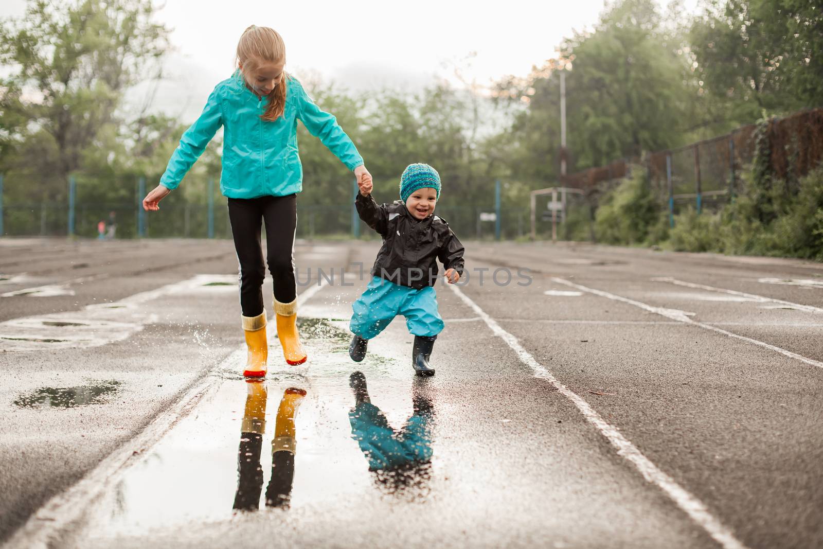 Boy and girl jumping in puddle in waterproof coat and rubber boots by Angel_a